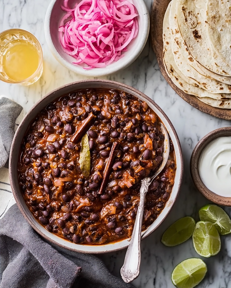 A bowl filled with black beans cooked in a rich, chunky tomato sauce with visible pieces of onion and celery, topped with a dark bay leaf and a cinnamon stick nestled inside. Next to the bowl, there is a white bowl with bright pink pickled onion slices, a stack of folded lightly browned corn tortillas on a wooden board, and a white bowl of creamy white sauce with a spoon in it. Lime wedges are scattered around the wooden surface, with two small glasses of a light yellow drink nearby. The entire arrangement is on a white marbled textured surface with a beige and black striped cloth napkin beside the large bowl. Photo taken with an iphone --ar 4:5 --v 7