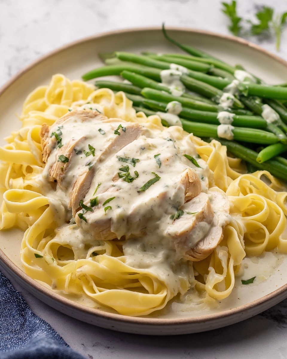 The dish shows three main layers on a white plate with a beige edge, placed on a white marbled surface. The bottom layer is light yellow fettuccine pasta arranged in a soft pile, showing a smooth texture with some strands looping around. On top of the pasta is a piece of light beige cooked chicken, sliced partially, covered with a creamy white sauce speckled with herbs. The sauce looks smooth and thick. To the side, there is a pile of bright green steamed green beans with the same white creamy sauce drizzled lightly over them. Small green herb pieces are sprinkled on the chicken and sauce for garnish. Photo taken with an iphone --ar 4:5 --v 7