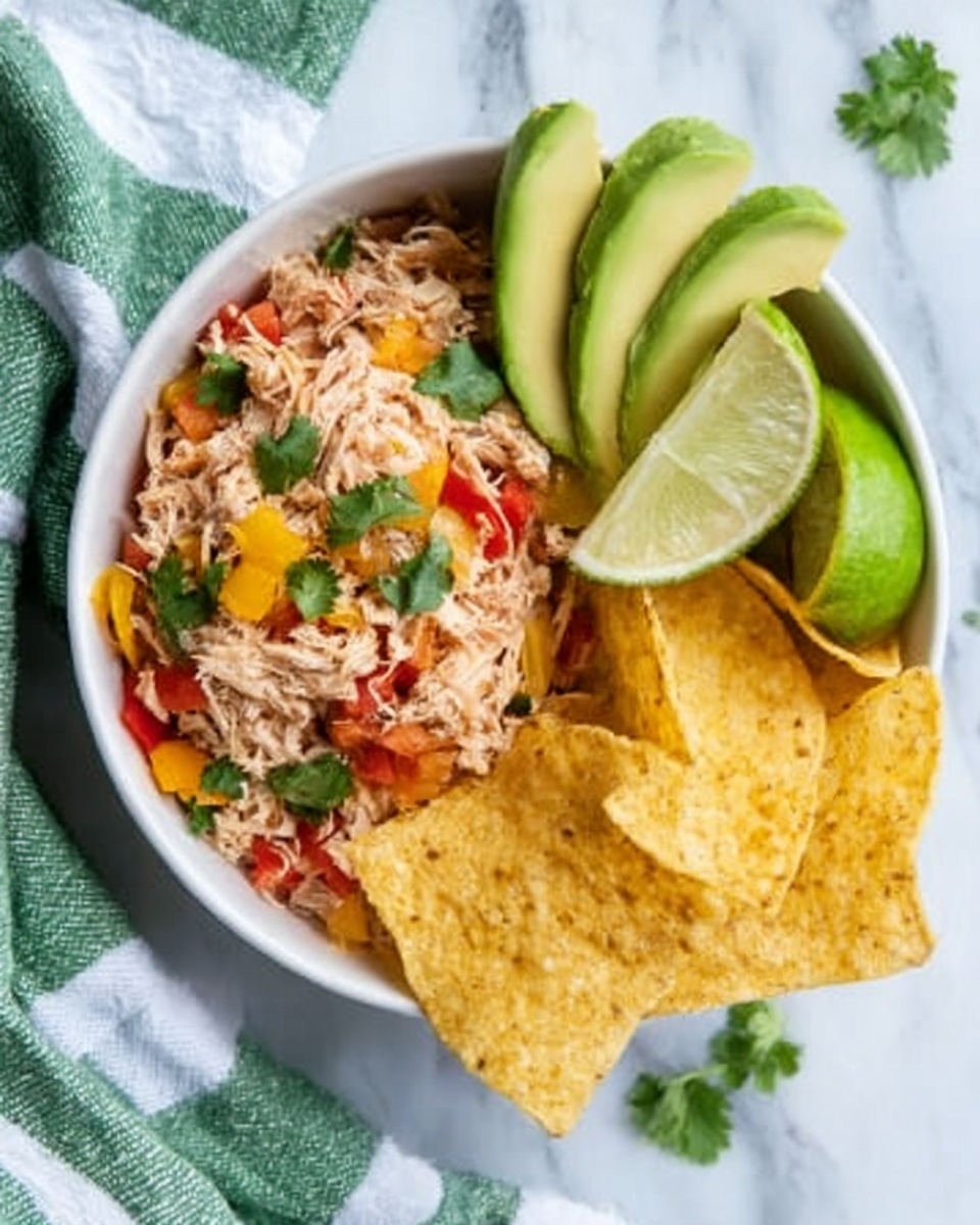 A white bowl holds three main layers: shredded light-colored chicken mixed with small pieces of red tomato and green herbs, positioned on one side; next to it, several golden yellow tortilla chips with a crisp texture; on the other side, neatly sliced avocado with a smooth green surface arranged in a fan shape. A wedge of lime rests near the chicken, and a few scattered cilantro leaves add green accents. The bowl is placed on a white marbled surface with a green and white striped cloth nearby. Photo taken with an iphone --ar 4:5 --v 7