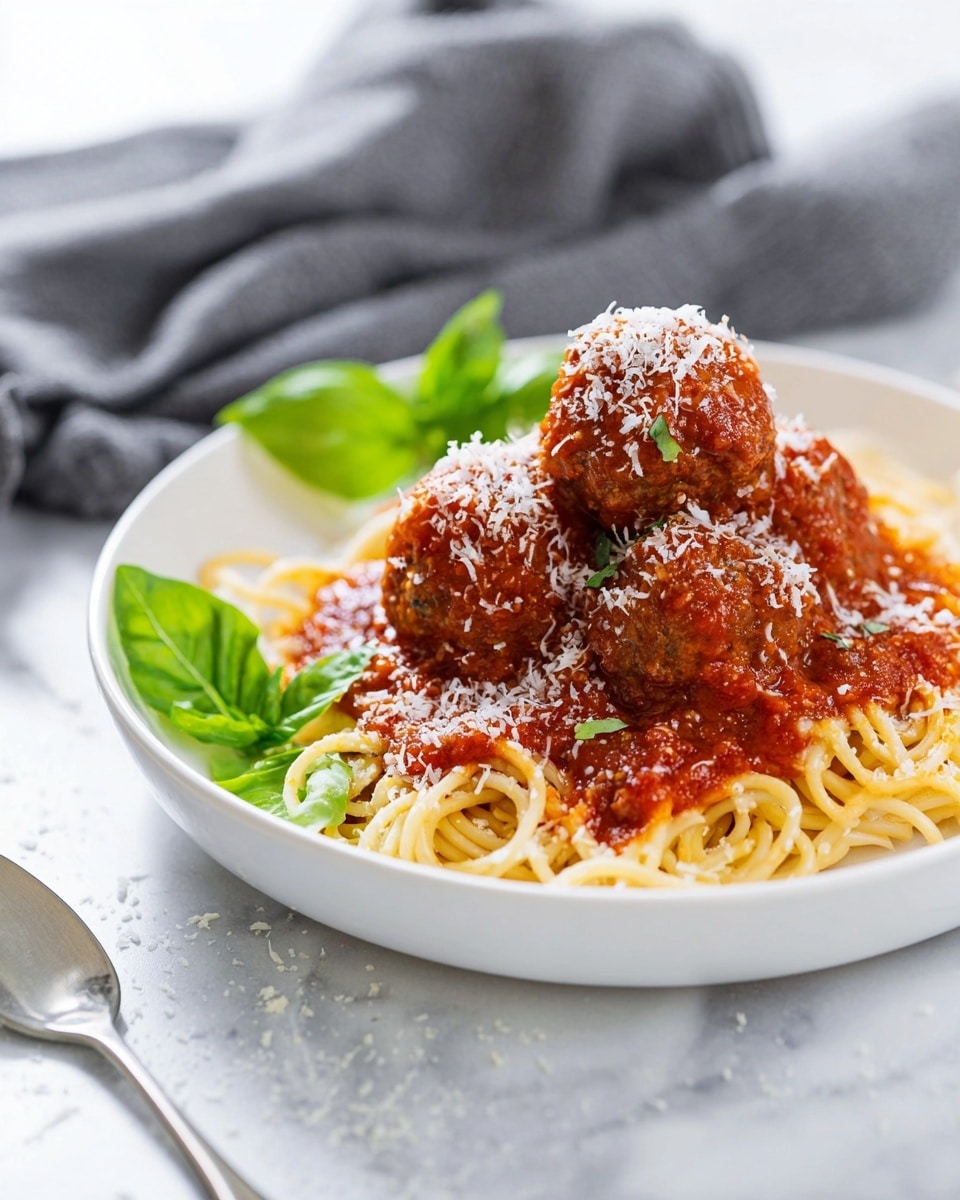 A white plate holds a bed of light yellow spaghetti noodles that are piled loosely at the base. On top, there are four large round meatballs coated evenly in a thick, rich red tomato sauce with visible herbs. Fresh green basil leaves are placed on one side of the meatballs, adding a pop of color. The dish is finished with finely grated white cheese scattered generously over the meatballs and sauce, giving a textured look. The plate sits on a white marbled surface, with a silver fork nearby. Photo taken with an iphone --ar 4:5 --v 7