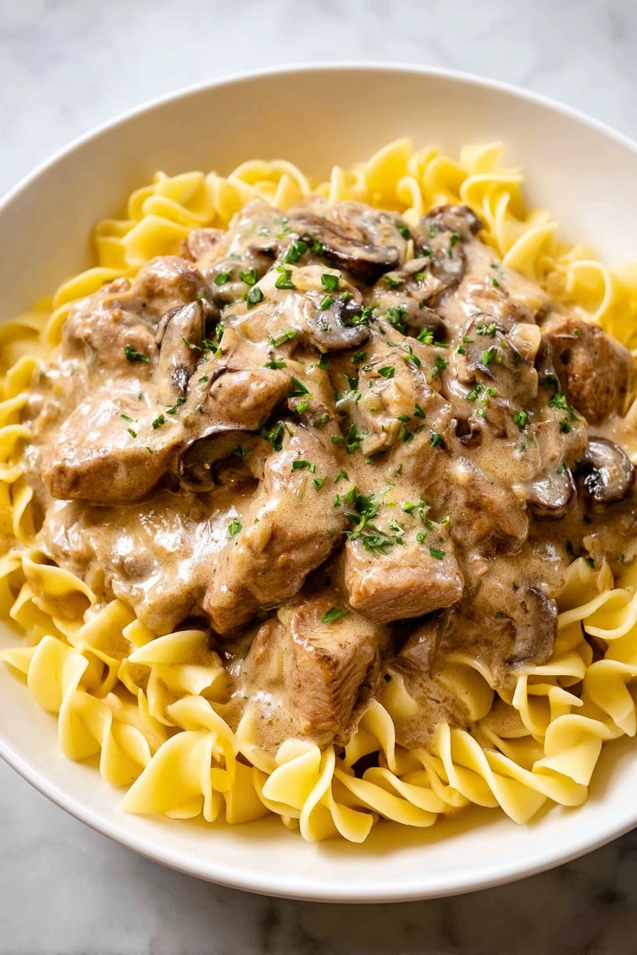 In a white bowl, there is a layer of yellow egg noodles spread evenly along the bottom and around the edges. On top, there is a thick layer of brown creamy sauce with visible pieces of cooked mushrooms and tender slices of meat covered in the sauce. Small green herbs are sprinkled on the sauce, adding a touch of color. The bowl is placed on a white marbled surface. photo taken with an iphone --ar 4:5 --v 7