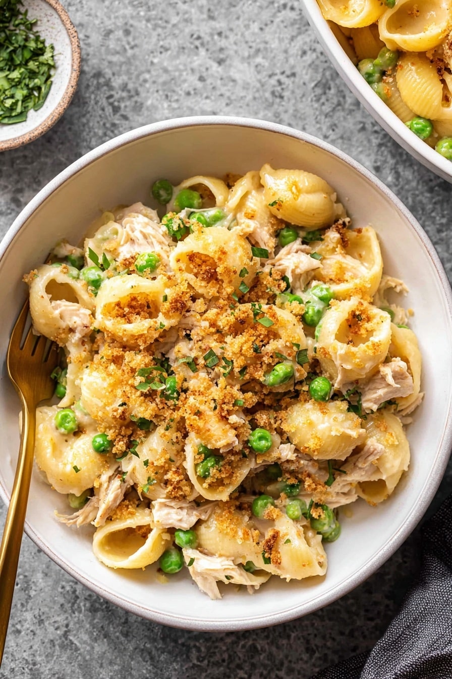 A white bowl with a brown rim holds a pasta dish made of short, round pasta shells mixed with small green peas and pieces of light brown cooked meat. The pasta is coated in a creamy, pale yellow sauce and topped with golden breadcrumbs. Small green herb leaves are scattered on top, adding color contrast. A gold fork rests inside the bowl, partially buried in the pasta. The bowl is placed on a white marbled surface, with a small white cloth visible on one side. Photo taken with an iphone --ar 4:5 --v 7