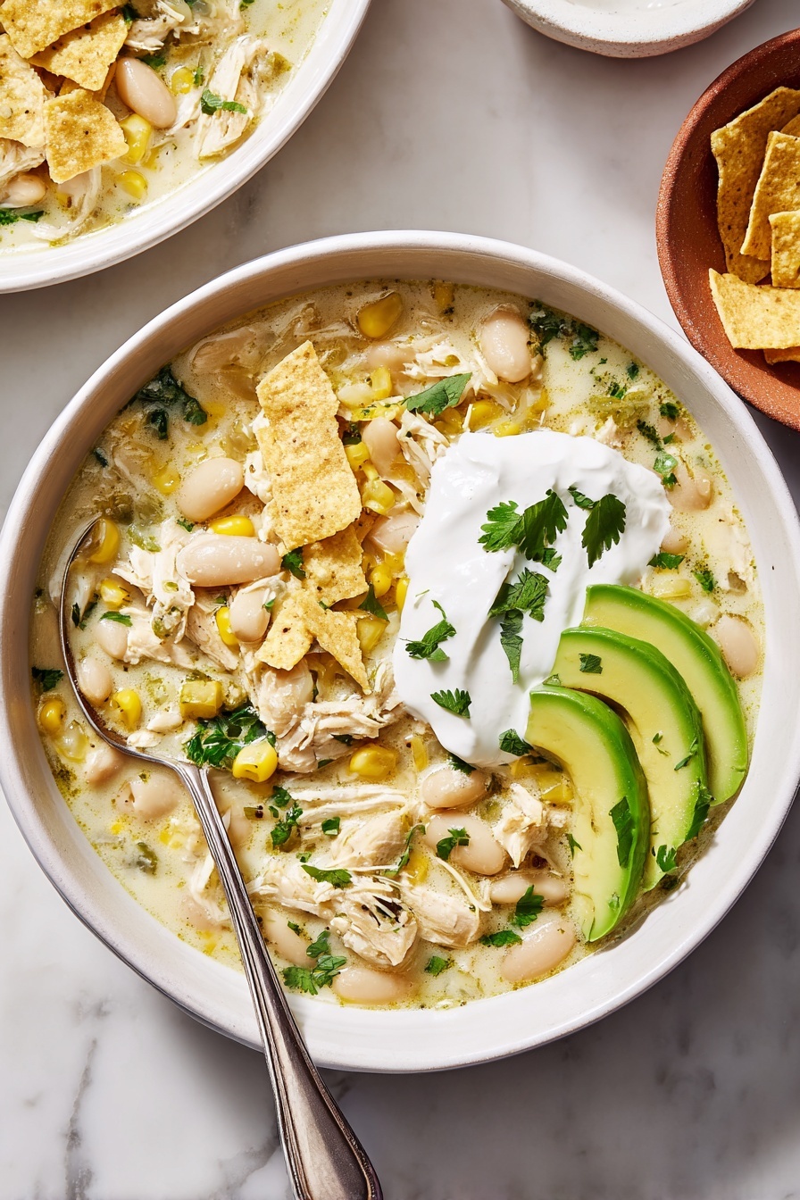 One-Pot White Chicken Chili Recipes Recipe 6 The image shows a white bowl filled with creamy soup containing shredded light-colored chicken, white beans, yellow corn, chopped green herbs, and pieces of crushed pale yellow tortilla chips mixed throughout. On one side of the soup, there is a dollop of smooth white sour cream topped with a few small green herb leaves. Next to it, three bright green slices of avocado are neatly placed on top of the soup, also garnished with small herb leaves. A silver spoon is resting inside the bowl on the left side. The bowl is set on a white marbled surface with part of another similar bowl visible at the top right corner and a glimpse of a white bowl of tortilla chips in the bottom right corner. Photo taken with an iphone --ar 4:5 --v 7