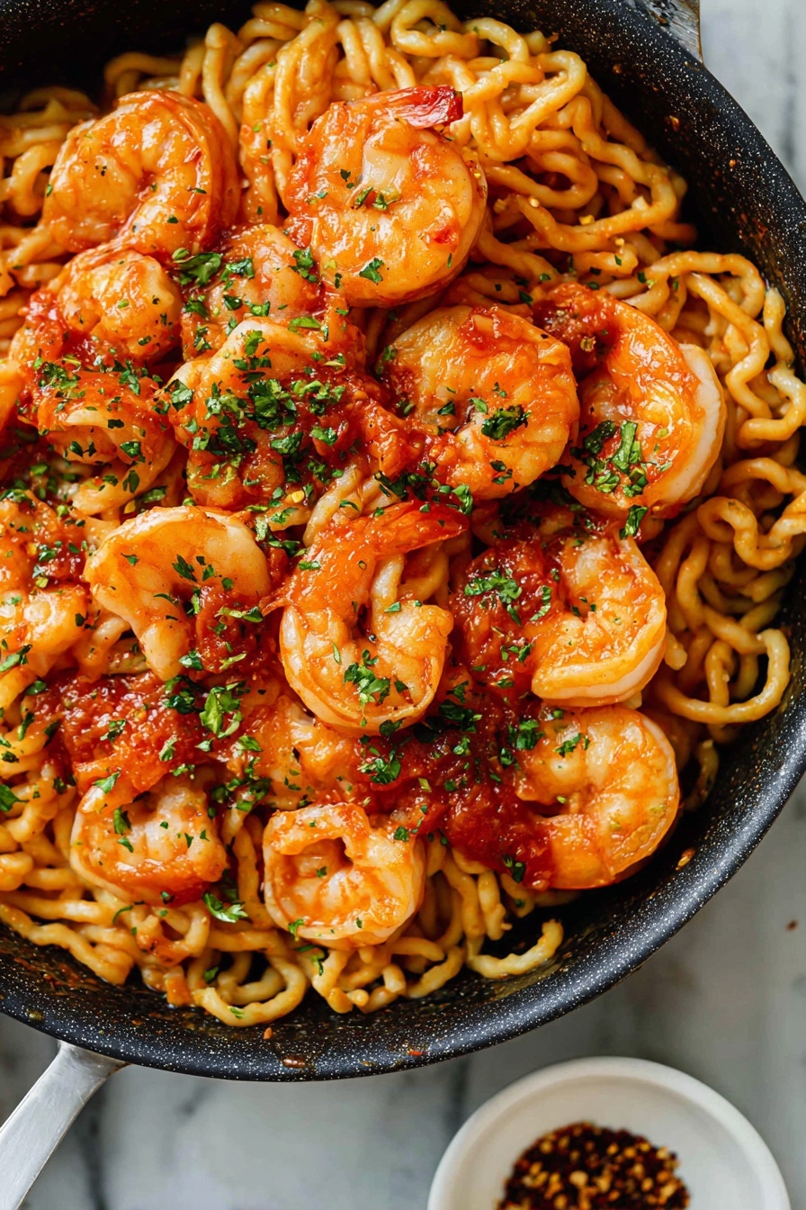A close-up view of a black pan filled with curled, pale yellow pasta mixed with bright orange cooked shrimp, each shrimp having a light red sauce and green herb bits sprinkled on top. The pasta has a wavy texture and sits beneath the plump shrimp, which have slightly charred spots adding texture. The dish is garnished with finely chopped green herbs scattered evenly across the shrimp and pasta. The pan rests on a white marbled surface, with a small white bowl containing red flakes placed nearby. photo taken with an iphone --ar 4:5 --v 7