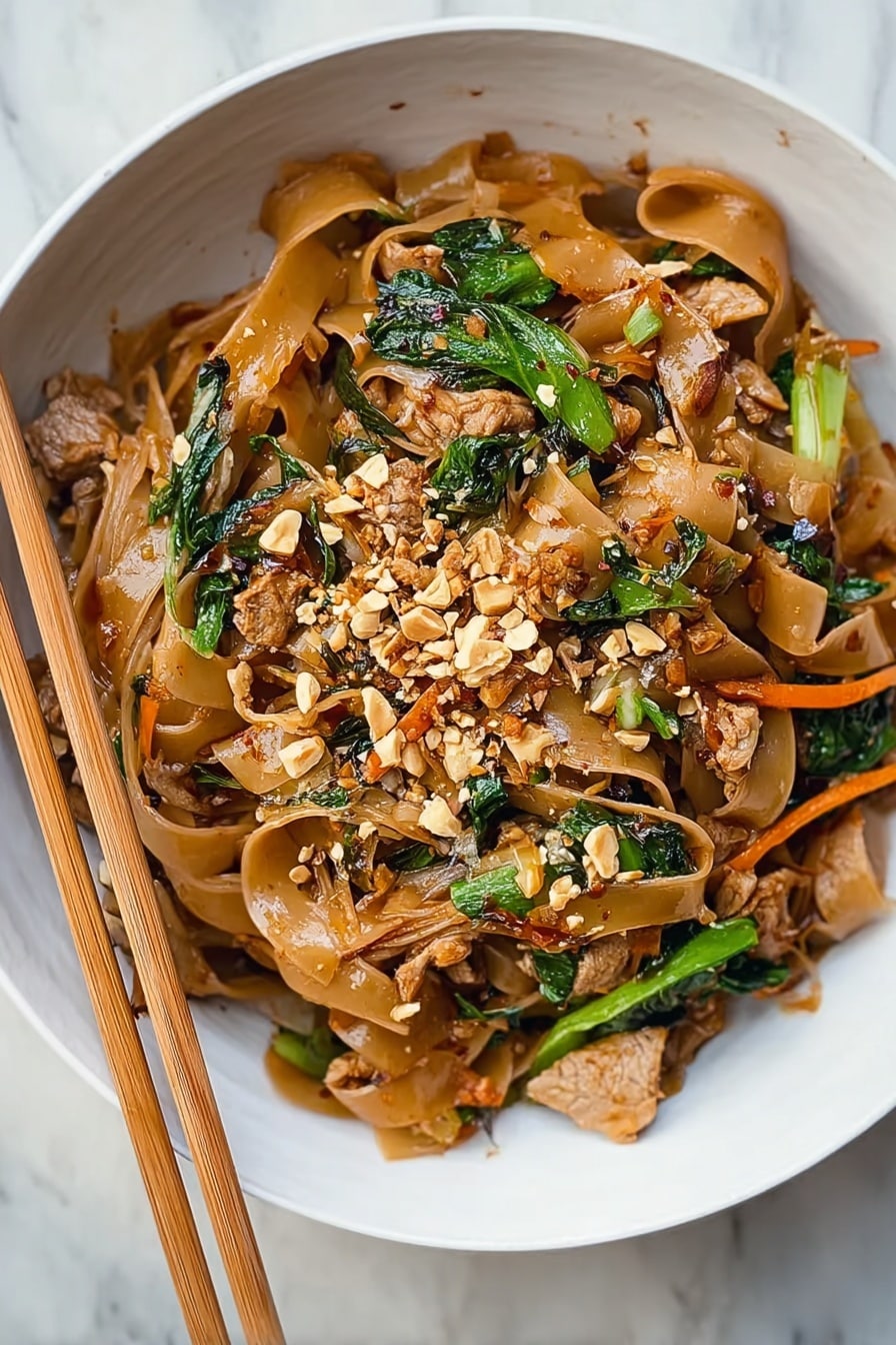 Easy Chicken Drunken Noodles Recipe 6 The dish shows a white bowl filled with wide, flat brown noodles mixed with cooked pieces of light brown meat, bright green leafy vegetables, and thin orange carrot strips. On top, small chopped peanuts are scattered, adding texture and light cream color. The noodles and vegetables are glossy, indicating a sauce coating. Light-colored chopsticks rest on the edge of the bowl, placed over a white marbled surface. Photo taken with an iphone --ar 4:5 --v 7