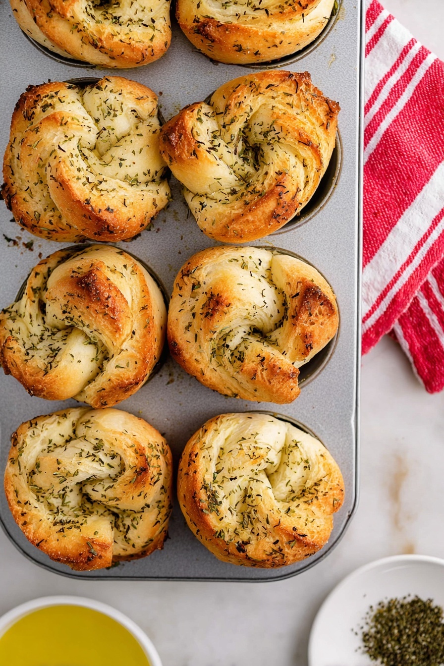 The image shows a metal muffin tray filled with golden brown biscuit knots. Each biscuit has several layers visible, with a light, fluffy inside and a crispy, slightly darker outside sprinkled with green herbs. The knots have an irregular, rustic texture with folded dough creating a soft, pillowy look. The tray sits on a white marbled surface with a red and white striped cloth on the right side. Some dried herbs are scattered around the base of the tray. Photo taken with an iphone --ar 4:5 --v 7