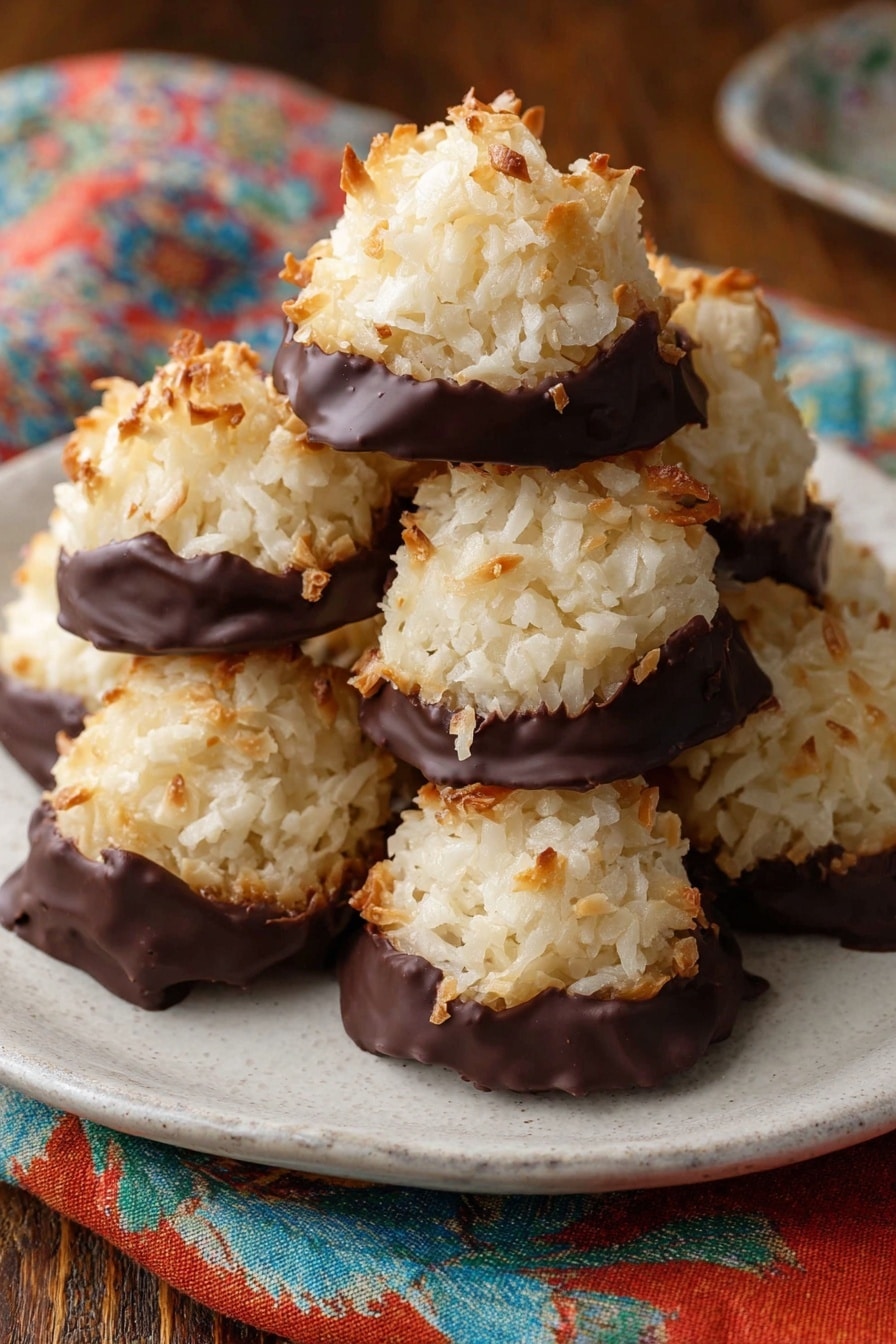 Chocolate Dipped Coconut Macaroons Recipe 6 The image shows six toasted coconut macaroons on a white parchment-lined baking tray. Each macaroon has two layers: a bottom layer of shiny dark chocolate that slightly spreads out unevenly on the parchment and a top dome-shaped layer of shredded, golden-brown toasted coconut with a rough, flaky texture. The macaroons are spaced evenly, and the baking tray has a worn, metallic look. The background includes a fabric with red, green, and yellow floral patterns. photo taken with an iphone --ar 4:5 --v 7