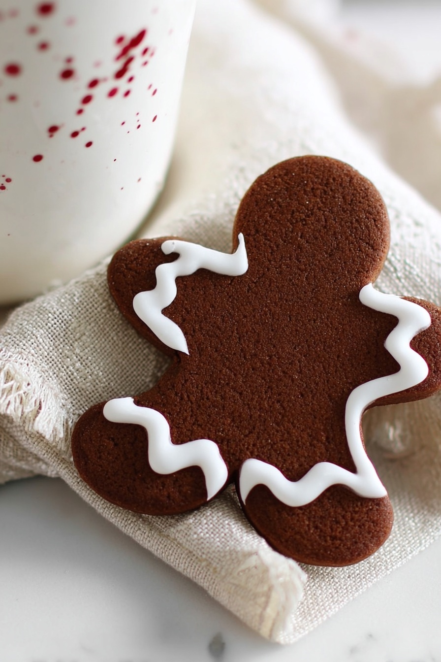 The image shows a single gingerbread cookie shaped like a person with a bite taken out of the upper left side. The cookie has a rich brown color with a slightly rough texture. Around its edge, there is one layer of smooth white icing that follows the cookie’s outline in a wavy line. The cookie rests on a white cloth with soft folds, and behind it, there is a white cup partially visible. The whole scene is set on a white marbled surface, giving a clean and bright background. photo taken with an iphone --ar 4:5 --v 7