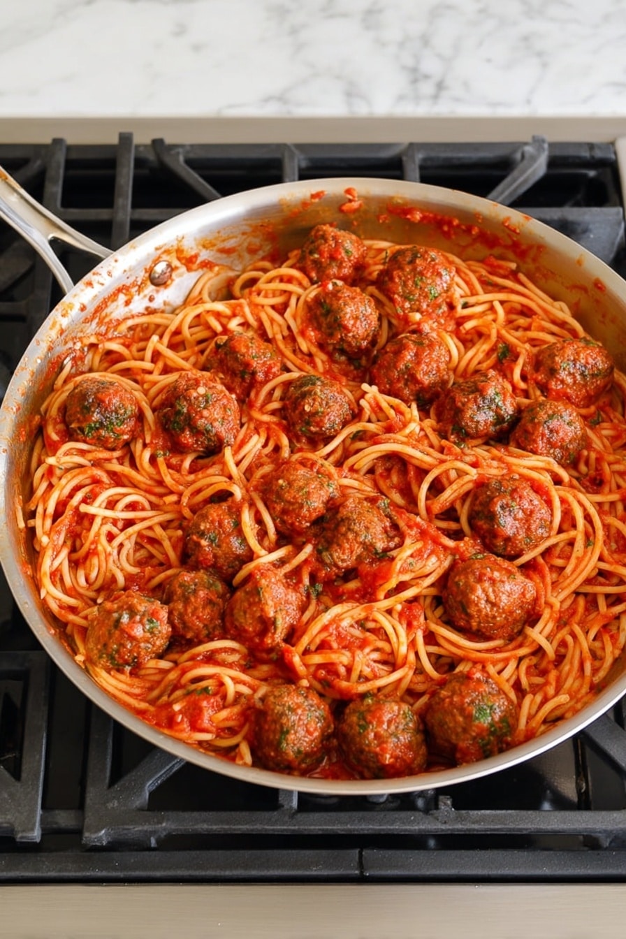 Easy Baked Meatballs with Spaghetti Recipe 5 A large metal pan filled with spaghetti pasta mixed with red tomato sauce and several round meatballs evenly spread on top and through the noodles. The spaghetti is coated with a rich, smooth sauce and looks soft. The meatballs have a brown color with green herbs mixed inside, showing a textured surface. The pan is placed on a gas stove, surrounded by a white marbled counter. The scene has a simple kitchen setting with neutral tones. photo taken with an iphone --ar 4:5 --v 7