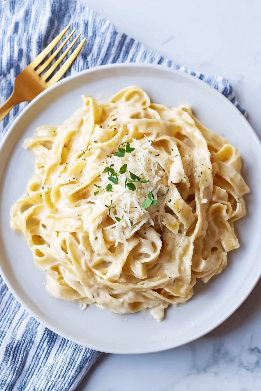 Creamy Fettuccine Alfredo Recipe 6 A white bowl filled with fettuccine pasta coated in a creamy, light beige sauce with a smooth texture. On top of the pasta, there is a small pile of finely shredded white cheese and a sprinkle of chopped green herbs. The bowl is placed on a white marbled surface, next to a blue and white striped cloth napkin with a shiny gold fork resting on it. The photo taken with an iphone --ar 4:5 --v 7