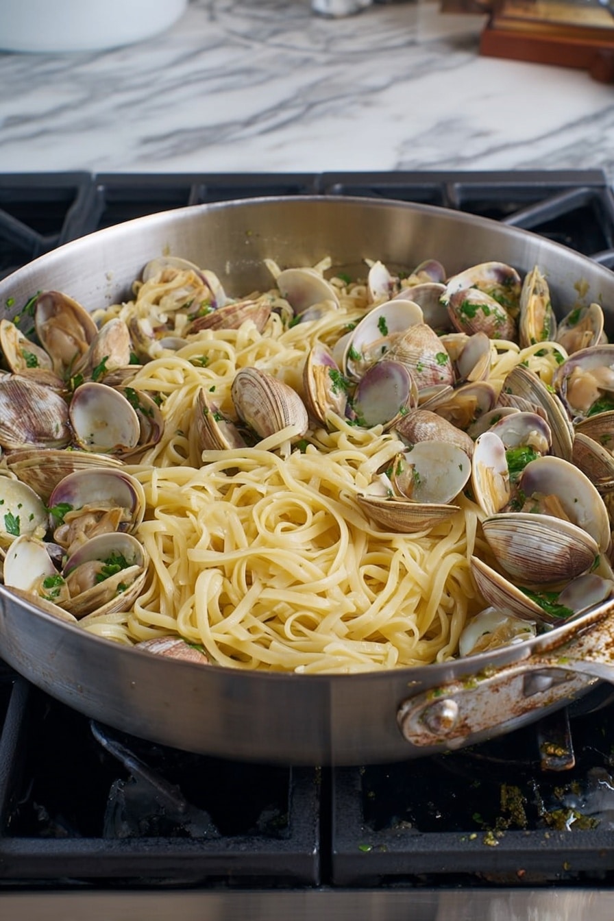 A large silver pan filled with a mix of long, light yellow pasta noodles and many clam shells in shades of light brown and gray, scattered evenly throughout the dish. Small green herbs are sprinkled on top and mixed in, along with tiny bits of chopped garlic and red chili flakes. The pan sits on a stovetop with black grates, and the background is a white marbled surface. The food looks fresh and ready to serve photo taken with an iphone --ar 4:5 --v 7