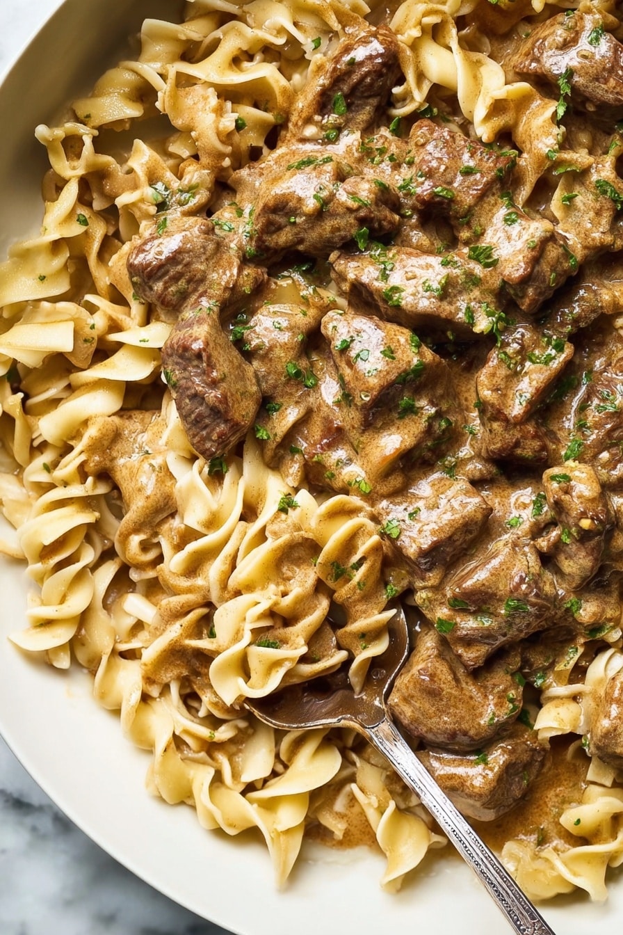 The image shows a close-up of a dish with thick, curly egg noodles in a light brown creamy sauce, mixed with several pieces of browned, tender meat strips. The noodles have a slightly curled texture and are coated evenly in the sauce, giving them a glossy look. The meat pieces are scattered throughout, with a few slices having seared marks and coated in sauce as well. Small green herb bits are sprinkled over the dish, adding a fresh contrast to the warm tones. The dish is served in a white bowl on a white marbled surface, with a metal spoon partially dipped inside the food, showing some sauce on it. photo taken with an iphone --ar 4:5 --v 7