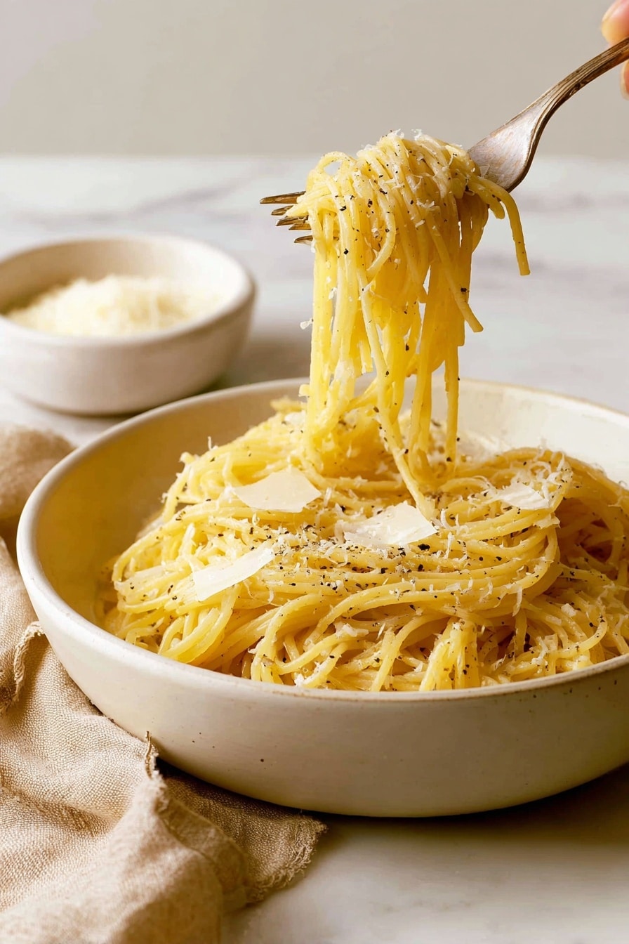 A close-up view of a white bowl filled with a neatly twirled mound of spaghetti pasta, coated lightly in a pale, creamy sauce. The pasta strands have a smooth texture and are sprinkled with grated white cheese and small bits of cracked black pepper, giving a light contrast throughout. To the left side of the bowl, a fork and a spoon rest casually on the edge, both with a metallic shine. The bowl sits on a white marbled surface. photo taken with an iphone --ar 4:5 --v 7