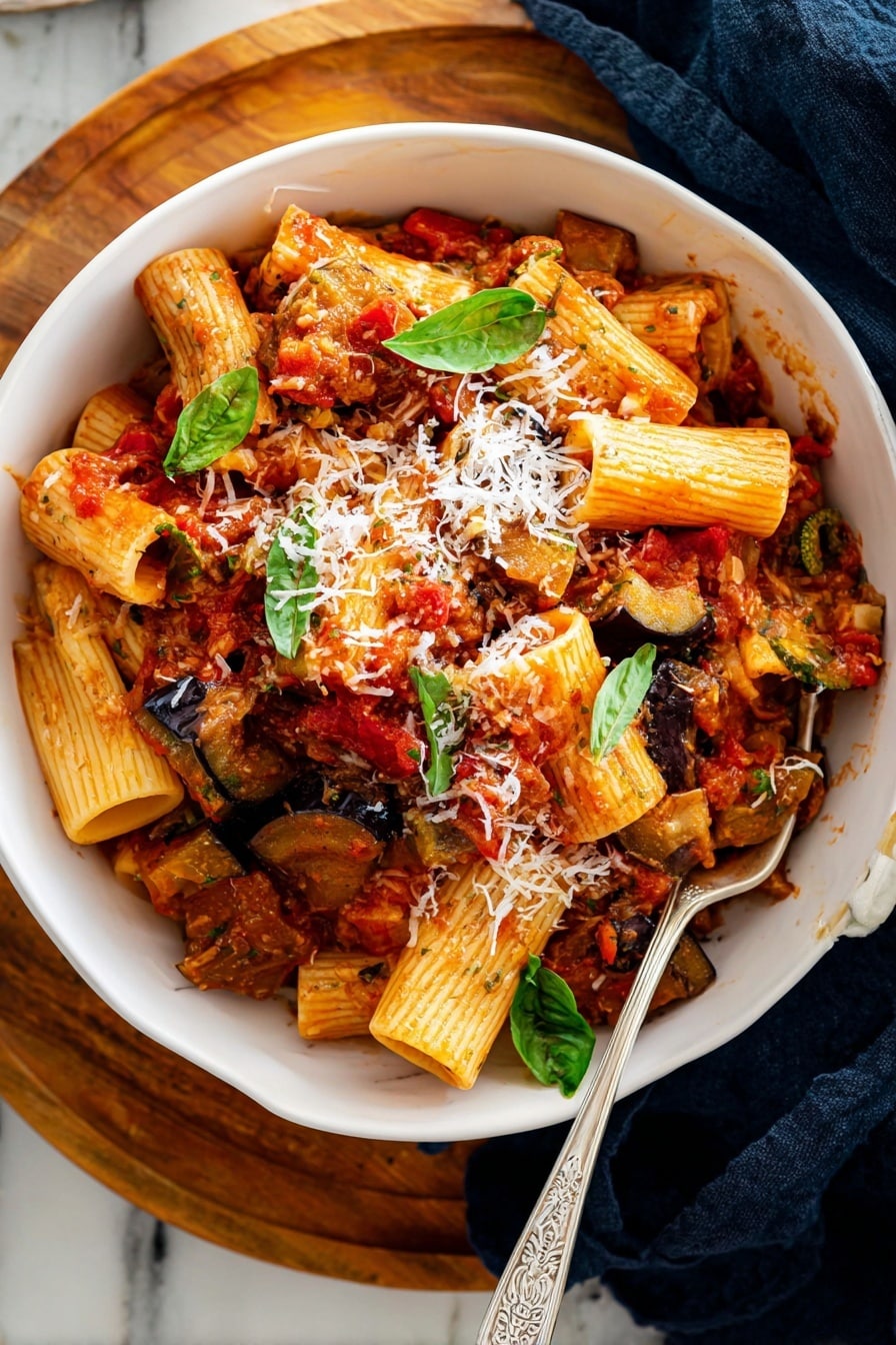 This image shows a close-up of cooked rigatoni pasta mixed with a thick textured red tomato sauce, pieces of soft cooked eggplant with dark purple skin, and sprinkled with green fresh basil leaves and shredded pale yellow cheese. The pasta is placed inside a black pan with a wooden spoon resting on the right side, partially lifting some pasta and vegetables. The background is a white marbled texture. photo taken with an iphone --ar 4:5 --v 7