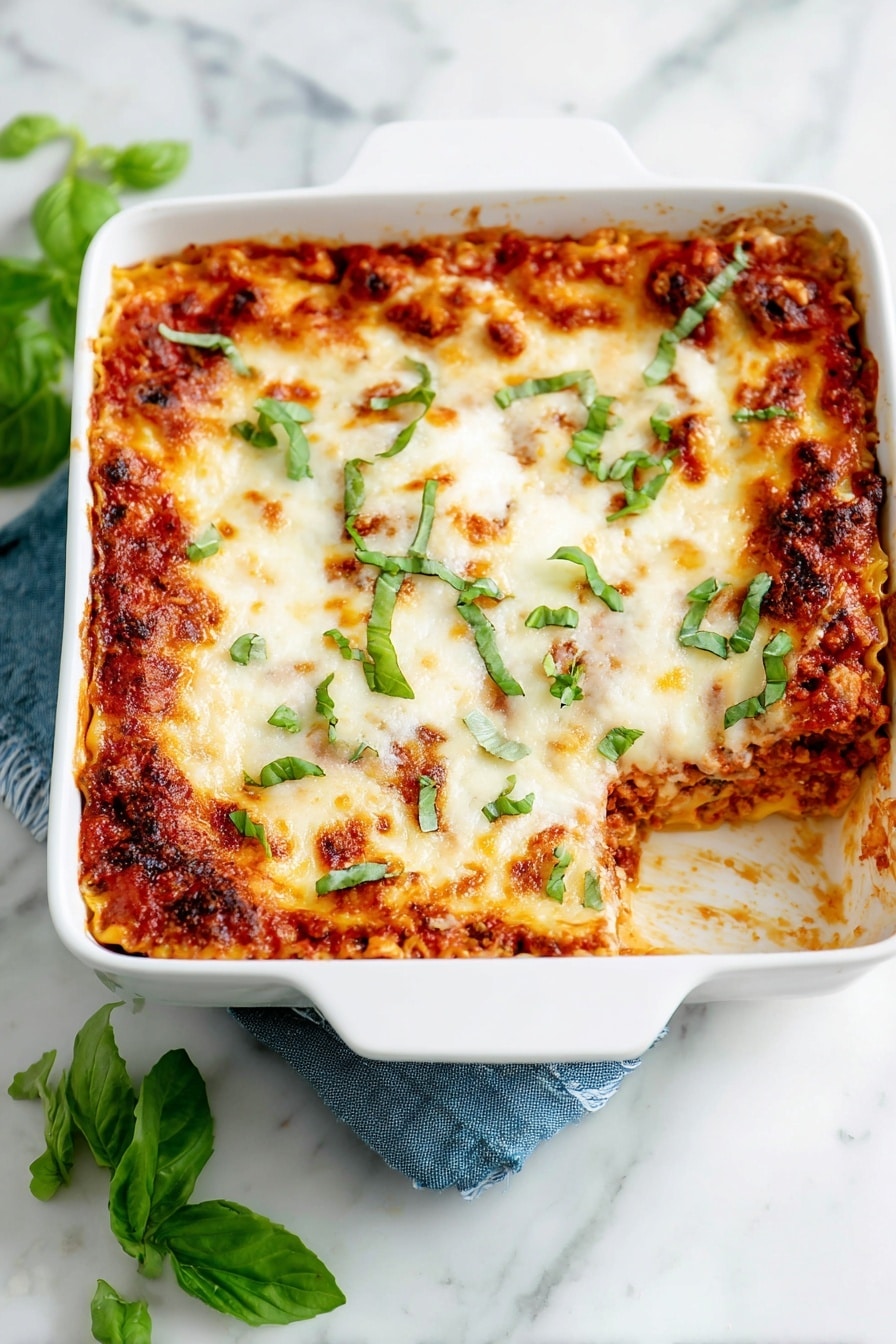 A white square baking dish holds a layered lasagna with visible layers of reddish tomato sauce and melted, slightly browned cheese on top. The cheese layer is smooth with some small bubbles and golden spots, sprinkled with fresh green basil leaves scattered unevenly across the surface. The sides of the dish show thick layers of sauce and pasta peeking through. The dish rests on a white marbled surface with a dark blue cloth partially under one handle, and some fresh basil leaves are placed nearby. Photo taken with an iphone --ar 4:5 --v 7