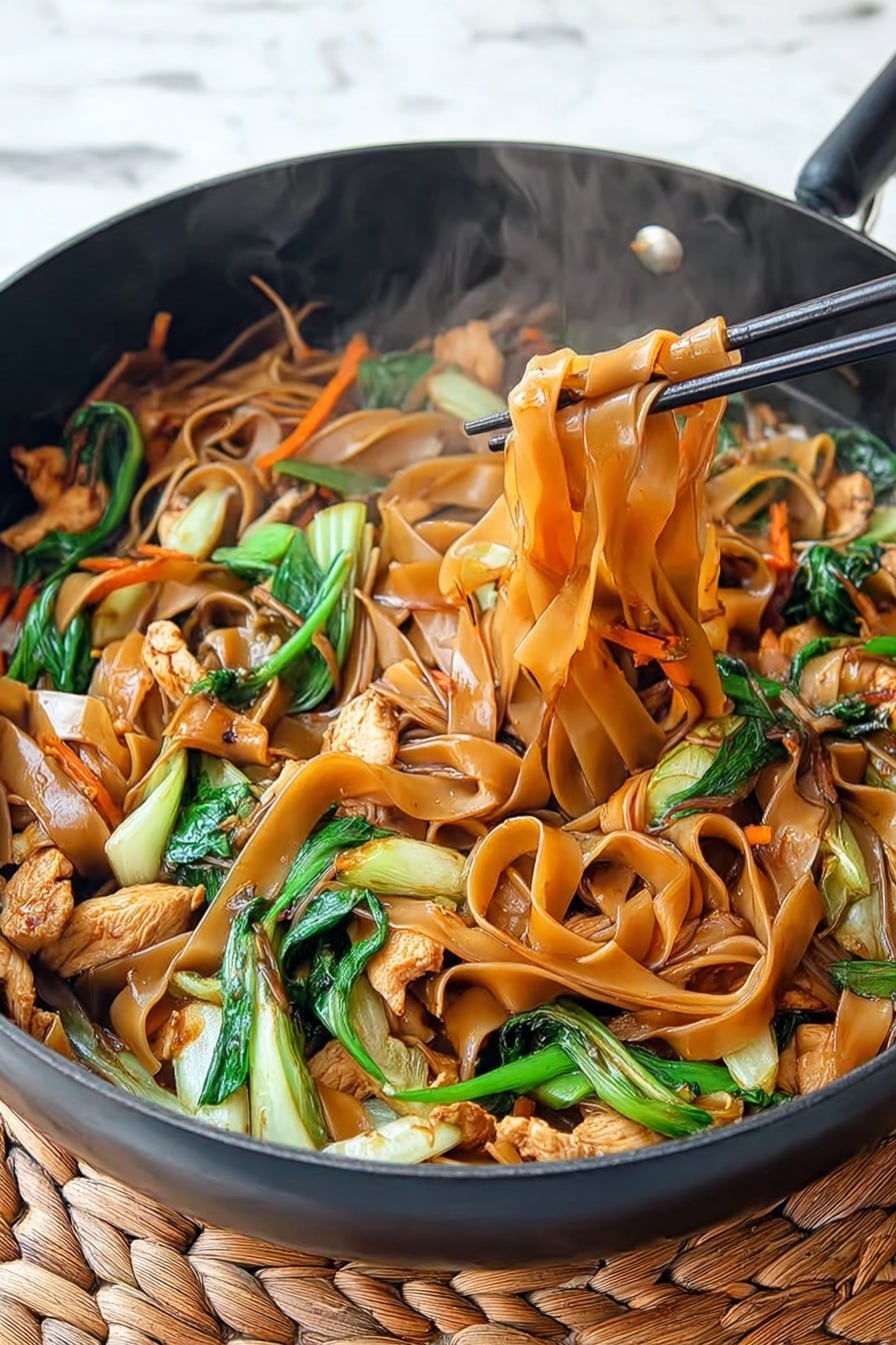 Easy Chicken Drunken Noodles Recipe 5 The image shows a black pan filled with flat, wide noodles that are light brown and glossy, mixed with pieces of light tan cooked chicken and bright green bok choy stalks and leaves. Thin strips of orange carrots are spread through the dish, adding color contrast. The noodles curl and twist around the other ingredients with steam rising, showing the dish is hot. A pair of black chopsticks is lifting some noodles and chicken from the pan. The pan sits on a woven beige mat on a white marbled surface. Photo taken with an iphone --ar 4:5 --v 7