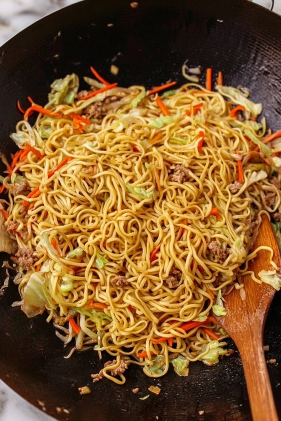 Easy Chicken Yakisoba Stir-Fry Recipe 5 The image shows a large black wok filled with cooked instant noodles mixed with thin strips of orange carrots, green cabbage, and small pieces of cooked meat scattered evenly throughout. The noodles have a glossy texture with a light brown color from the sauce they are cooked in, and the vegetables add some fresh color contrast. A wooden spoon is partially visible on the right side of the wok, resting on the noodles. The wok surface looks clean and slightly shiny, making the food appear hot and fresh. The background is a white marbled texture. photo taken with an iphone --ar 4:5 --v 7