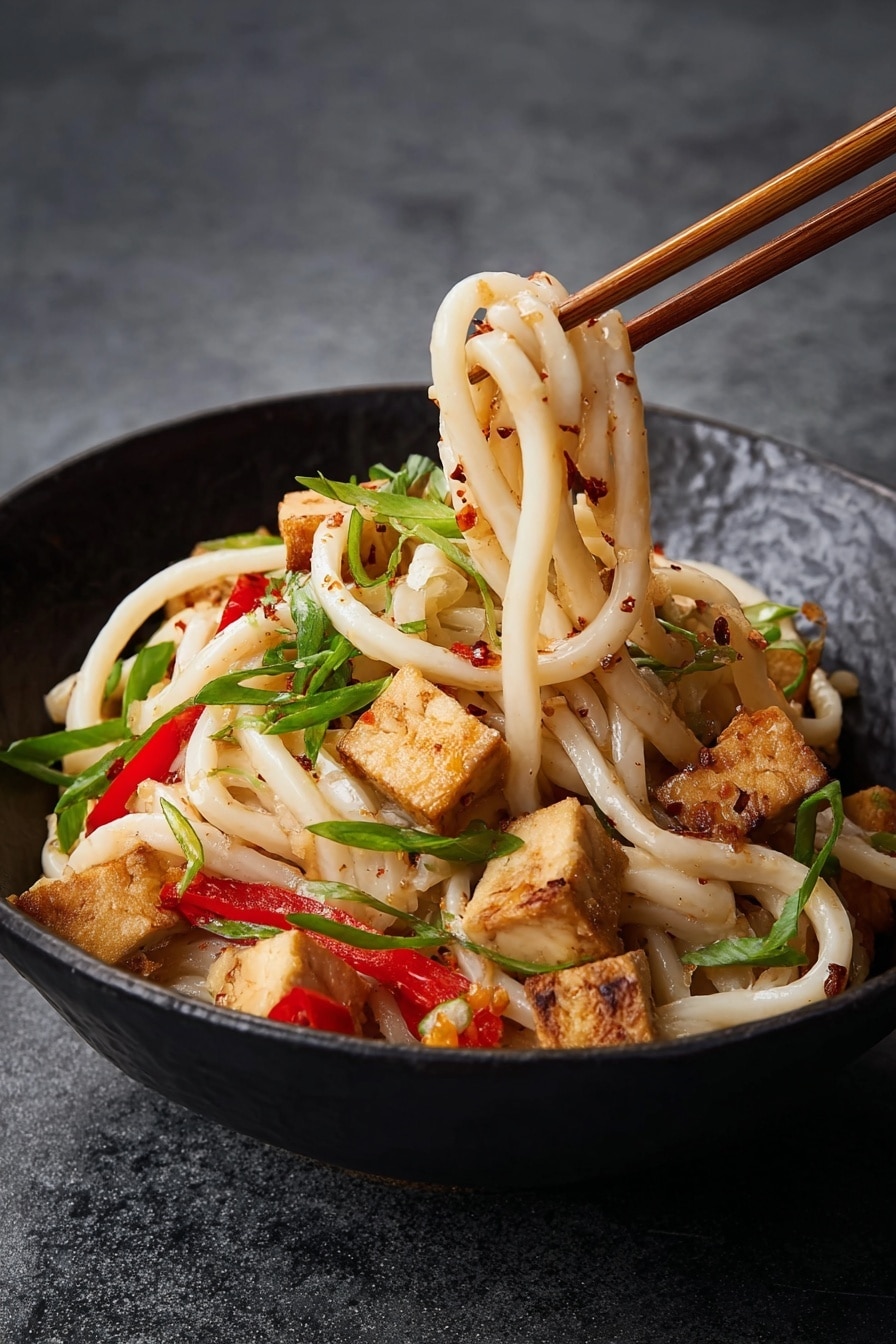 Garlic Peanut Noodles Recipe 6 A bowl holds a dish with thick, white noodles as the bottom layer, mixed with light brown fried tofu cubes scattered on top, showing a crispy texture. There are thin strips of bright red bell pepper interspersed between the noodles and tofu, along with small pieces of light tan peanuts. Thin, curly green onion slices are spread across the dish, adding a fresh vivid green color. The dish has little dark brown chili flakes or sauce drops sprinkled throughout, giving a spicy touch to the otherwise soft and chewy textures. The bowl is white and sits on a smooth white marbled surface. photo taken with an iphone --ar 4:5 --v 7