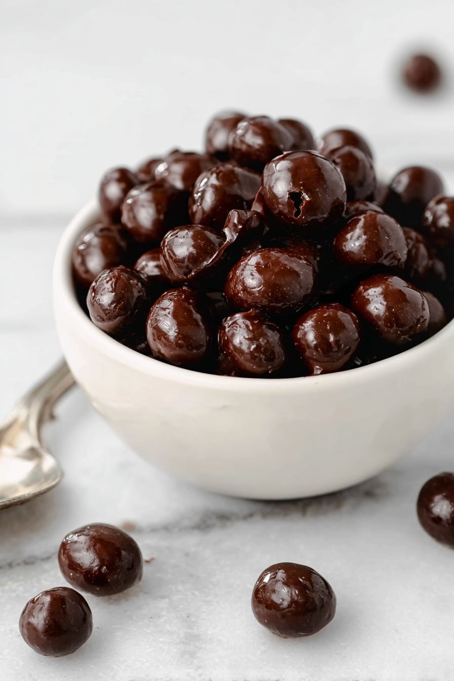 A small white bowl filled with shiny, dark brown chocolate-coated nuts piled high inside. The nuts have a smooth and glossy texture with some cracks showing their natural shape. Around the bowl on a white marbled surface, a few more chocolate-coated nuts are scattered, with some resting on a silver spoon at the bottom left corner. Photo taken with an iphone --ar 2:3 --v 7 - Chocolate Covered Espresso Beans, espresso beans snack, homemade coffee candies, caffeine chocolate treats, quick coffee chocolate recipe