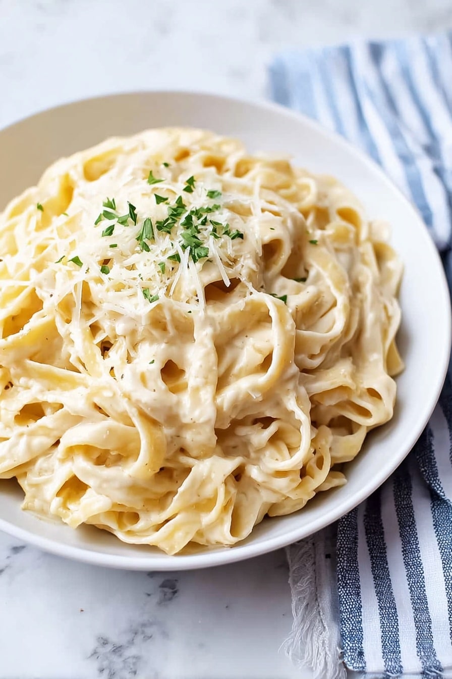 Creamy Fettuccine Alfredo Recipe 5 The image shows a white plate full of creamy fettuccine pasta coated in a smooth, light-colored sauce. The soft, flat pasta ribbons are layered evenly and topped with a small pile of grated cheese and chopped green herbs placed in the center. The plate rests on a white marbled surface with a folded blue and white striped cloth nearby. The overall appearance is simple and fresh. photo taken with an iphone --ar 4:5 --v 7