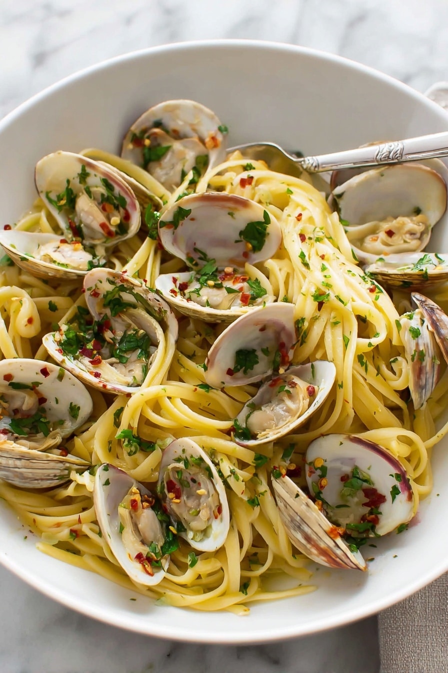A white bowl filled with a layer of light yellow spaghetti noodles mixed with small pieces of green herbs and bits of red chili. On top and around the noodles, open clams with white and gray shells reveal tender beige clam meat inside. The noodles and clams are coated lightly with oil and small bits of garlic. A silver fork rests inside the bowl on the right side, and the bowl sits on a white marbled surface with a glass of water blurred in the background. photo taken with an iphone --ar 4:5 --v 7