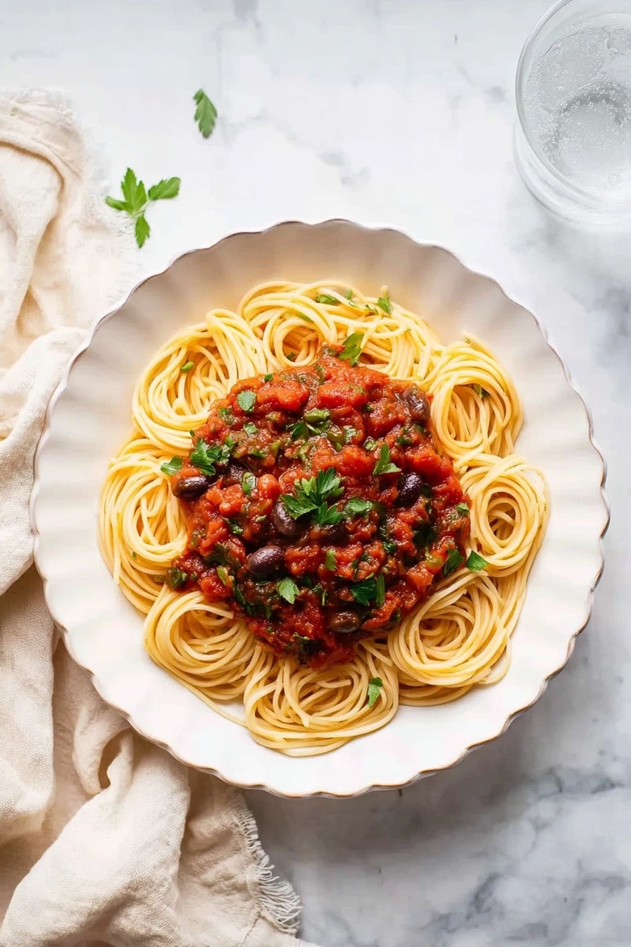 A white plate with a large serving of spaghetti noodles twisted into a loose nest in the center, topped with a red tomato sauce mixed with chunks of vegetables like mushrooms and bits of green herbs, giving the sauce a textured look. The sauce is spread over the top and tucked under some noodles, adding a mix of red, green, and brown colors that contrast with the light yellow pasta. A silver fork rests on the right side of the plate, partially twirling the noodles. The plate is set on a white marbled surface with a cream-colored cloth to the left, and a clear glass of water is placed on the upper right side near the plate. Photo taken with an iphone --ar 4:5 --v 7