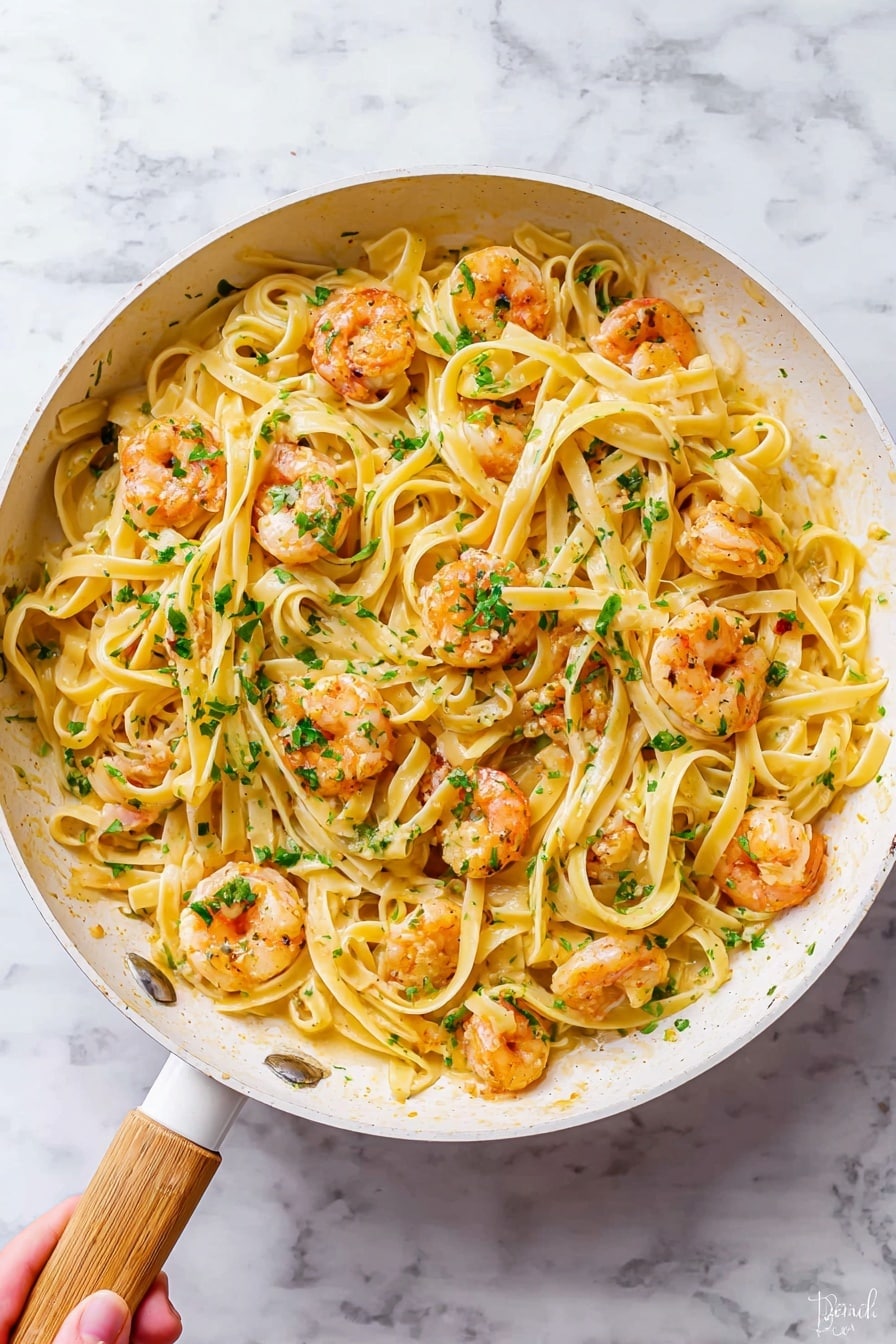 A white pan with a light wooden handle holds a dish of creamy fettuccine pasta mixed with cooked shrimp. The pasta strands are pale yellow, smooth, and twisted around the pinkish-orange shrimp. Small green parsley pieces are scattered evenly over the pasta and shrimp, adding bursts of color. The pan sits on a white marbled surface. photo taken with an iphone --ar 4:5 --v 7