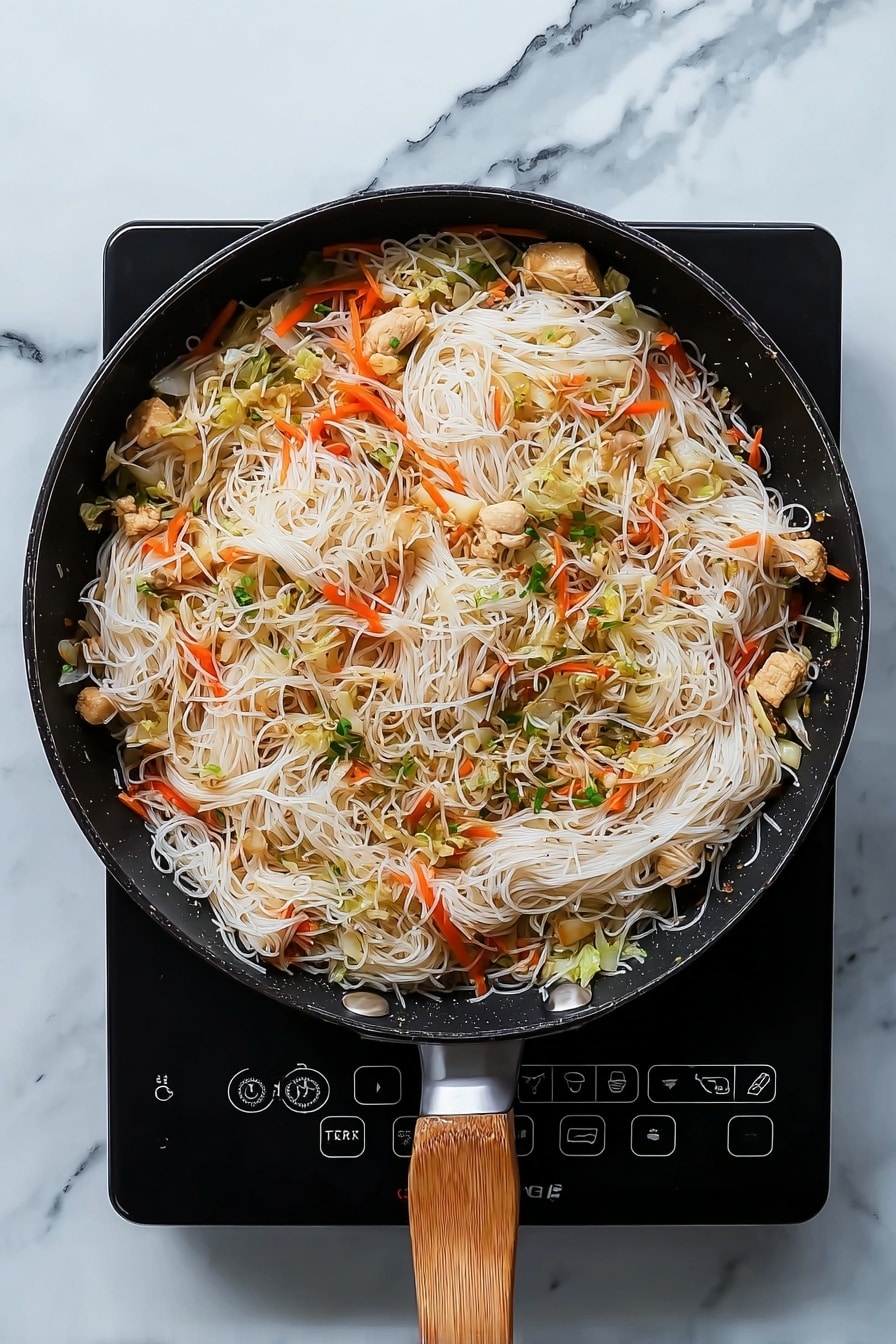 Easy Chicken Pancit Stir-Fry Recipe 5 A black pan with a wooden handle holds a dish of thin white rice noodles mixed with light brown cooked chicken pieces, thin orange carrot strips, and small pieces of pale green cabbage. The noodles form a soft, slightly tangled layer covering most of the pan's surface, with the vegetables and chicken spread evenly throughout. The pan sits on a white marbled countertop, which adds a clean, bright look to the scene. photo taken with an iphone --ar 4:5 --v 7