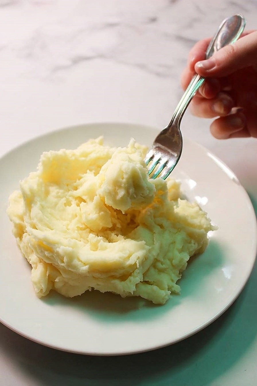 The image shows a white bowl filled with creamy mashed potatoes that are smooth with a few small lumps visible. A silver spoon is scooping some of the mashed potatoes from the bowl, resting partly inside it. The mashed potatoes have a pale yellow color with small brown specks scattered throughout. The bowl is placed on a white marbled surface, creating a clean and bright background. photo taken with an iphone --ar 4:5 --v 7