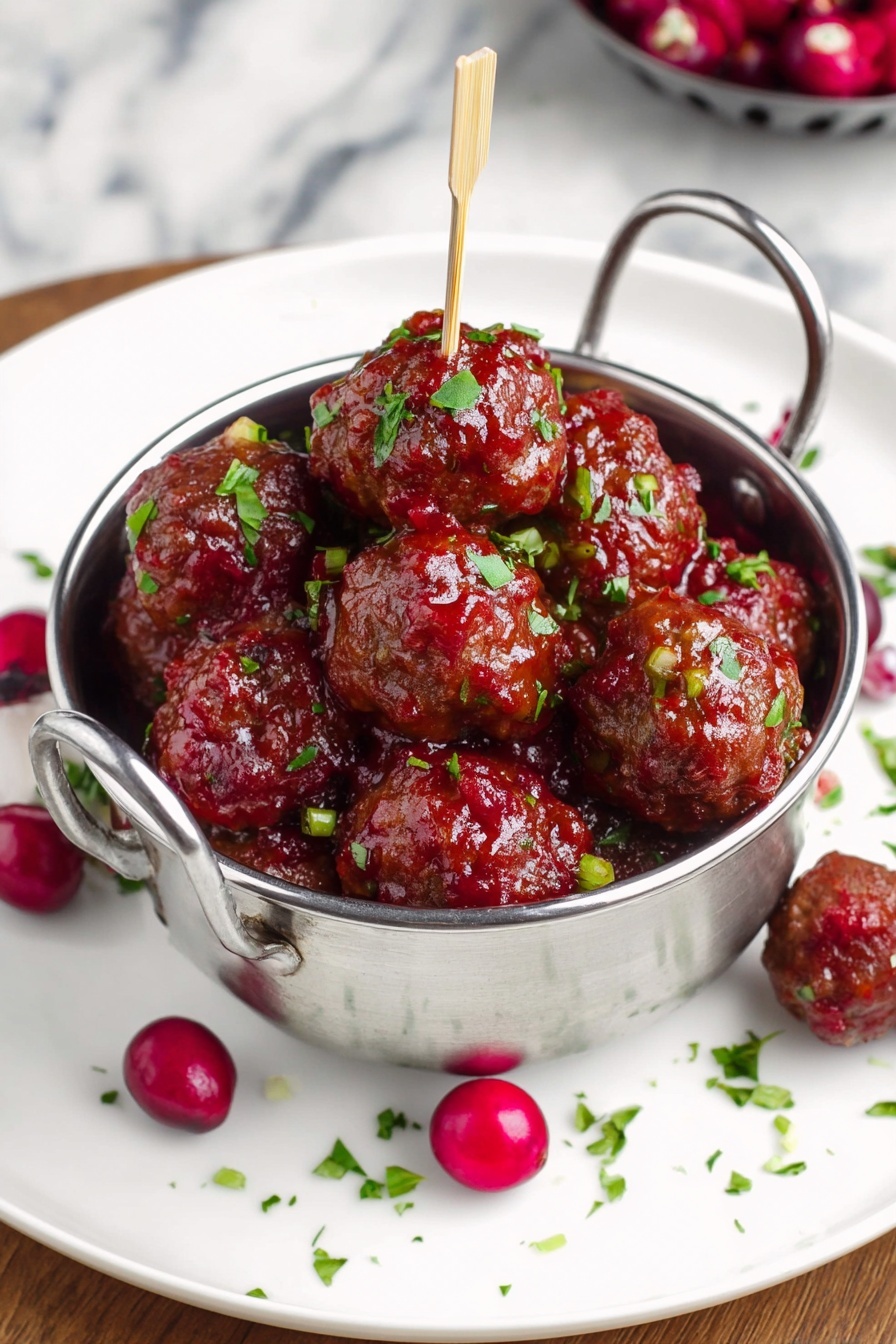A round silver bowl filled with about twenty small brown meatballs covered in a shiny red sauce with small green herb pieces scattered on top and mixed in. The meatballs sit closely together, and there are several whole red cranberries mixed in and placed around the bowl. The bowl is on a large white plate with a few more cranberries and herb bits scattered on it. A single meatball on a toothpick rests on the edge of the plate. The background surface is a white marbled texture with a small glass bowl of green herbs visible on the right. photo taken with an iphone --ar 4:5 --v 7