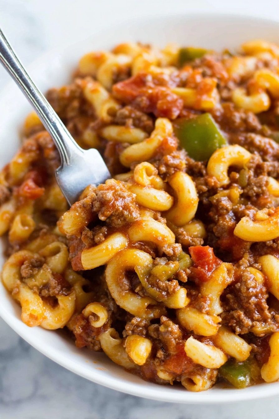 A close-up view of a white bowl filled with a mix of ground meat, elbow macaroni, diced green peppers, and small tomato chunks, all coated in a rich brownish-red sauce. The pasta is soft and curved, evenly mixed with the tender textured meat and vegetables. A silver fork is partially visible, resting on the edge of the bowl, subtly catching light. The whole dish is vibrant with warm tones and glossy textures, set against a white marbled background. Photo taken with an iphone --ar 4:5 --v 7