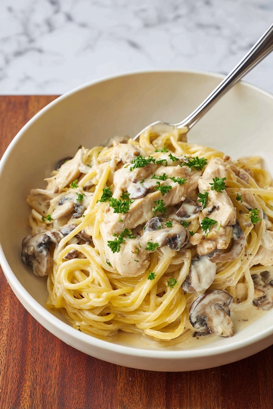 A white bowl holds a creamy pasta dish with two main layers; the bottom layer is a thick sauce mixed with pieces of white chicken and sliced light brown mushrooms, while the top layer is a nest of long, pale yellow spaghetti noodles. Small green parsley leaves are sprinkled on top, adding bright color to the dish. A fork rests on the right edge of the bowl, partially under the noodles. The bowl sits on a white marbled surface. photo taken with an iphone --ar 4:5 --v 7