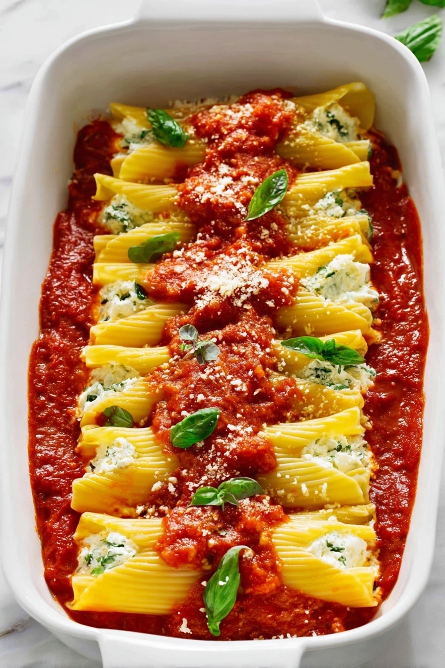 The image shows a white baking dish filled with a layer of deep red tomato sauce at the bottom, topped with two rows of large yellow pasta tubes filled with white cheese and green herbs inside. On top of each pasta layer, dollops of chunky red tomato sauce are spread unevenly. Fresh green basil leaves are scattered over the dish, along with light white shreds of cheese sprinkled on top. The dish sits on a white marbled surface. photo taken with an iphone --ar 4:5 --v 7
