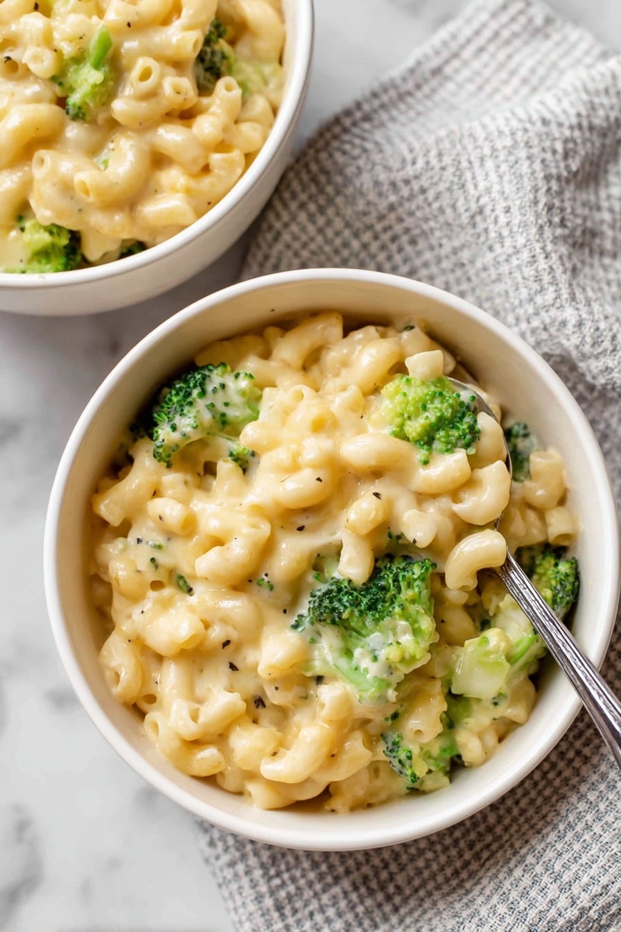 Broccoli Mac and Cheese Recipe 5 Two white bowls filled with creamy macaroni and cheese mixed with bright green broccoli pieces, showing a smooth, cheesy sauce covering the small, curved pasta. The bowls sit on a white marbled surface alongside a wrapped block of European style butter resting on a white cloth with a simple dark grid pattern. Loose dry macaroni noodles are scattered near the bowls, adding texture to the scene. Photo taken with an iphone --ar 4:5 --v 7