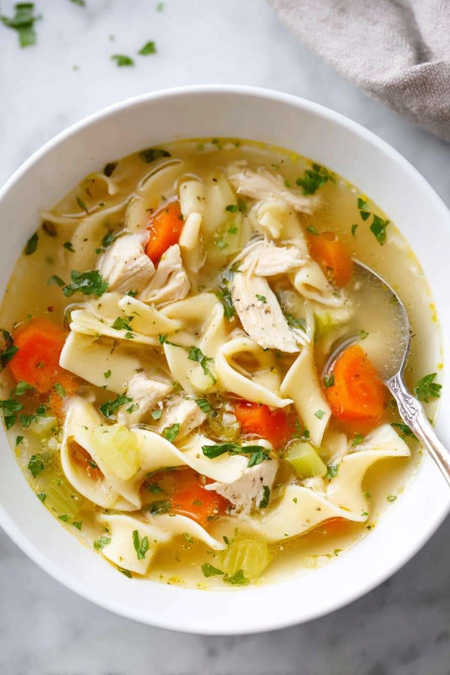 Homemade Chicken Noodle Soup Recipe 6 A white bowl filled with clear broth soup showing soft wide noodles folded gently and mixed with orange carrot slices and light green celery pieces. There are small light chicken pieces scattered throughout the soup, with green herbs sprinkled on top for color. The bowl sits on a white marbled surface with a spoon nearby. The soup looks warm and fresh with a mix of soft and slightly crunchy textures. photo taken with an iphone --ar 4:5 --v 7