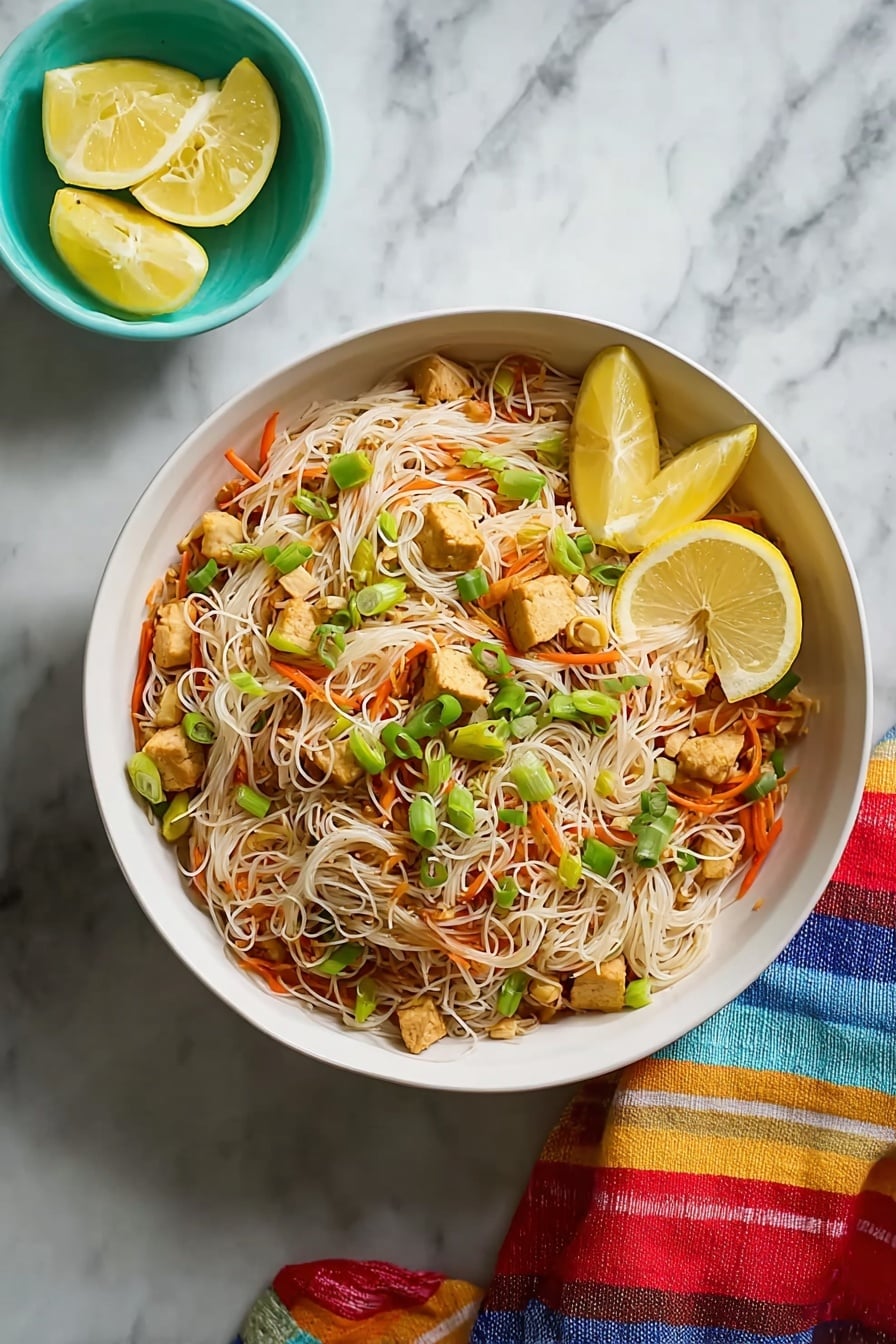 Easy Chicken Pancit Stir-Fry Recipe 6 A large white bowl filled with thin, light brown rice noodles mixed with small pieces of light brown tofu, thin orange carrot strips, and green vegetable bits scattered throughout. On top, green chopped scallions add a fresh touch. Two lemon wedges sit on the side inside the bowl. The bowl is on a white marbled surface, with a small turquoise bowl holding lemon wedges nearby, and a colorful striped cloth to the side. Photo taken with an iphone --ar 4:5 --v 7