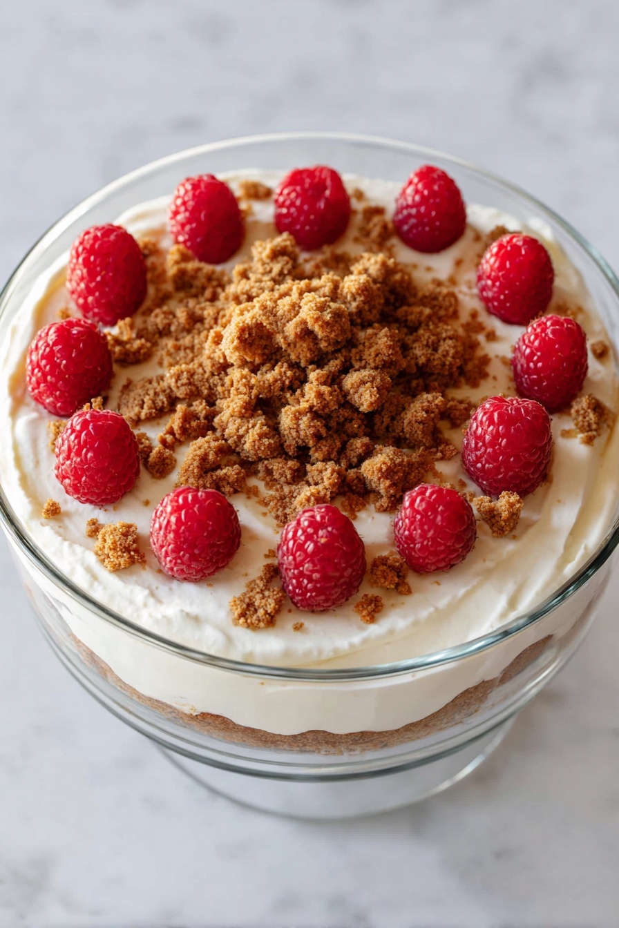 A clear glass bowl on a white marbled surface holds a creamy white dessert with three layers visible from the top: a smooth white whipped cream base, topped with scattered brown crumbly cookie bits, and bright red raspberries arranged in a circle near the edge and a smaller cluster in the center covered by more cookie crumbs. The whipped cream is softly textured with gentle swirls, and the crumbs add a rough contrast while the raspberries add a vibrant color pop. photo taken with an iphone --ar 4:5 --v 7