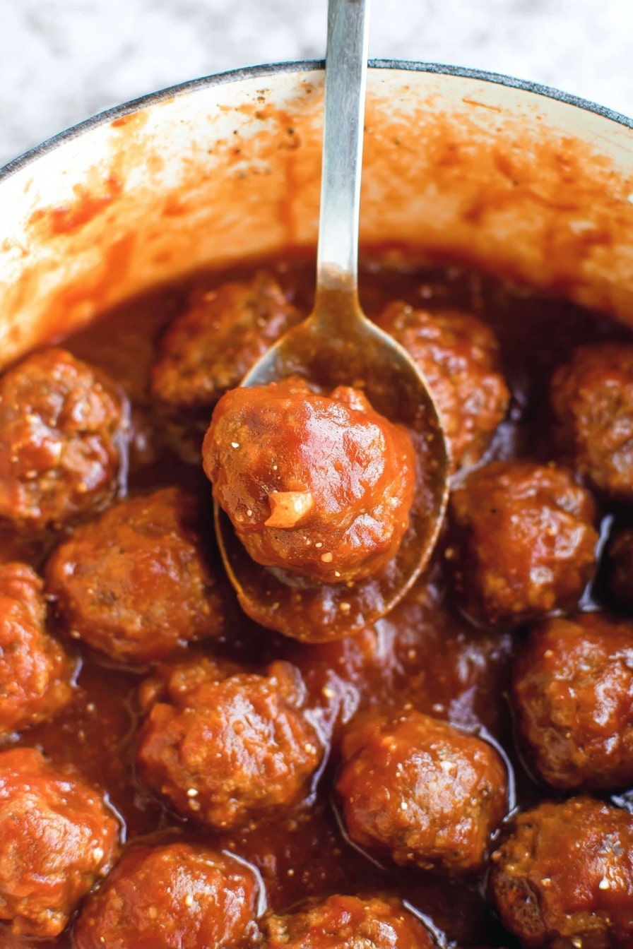This image shows many small, round meatballs covered in a thick reddish-brown sauce. The meatballs have a slightly shiny and moist texture, with some small bits of seasoning visible on their surface. They sit closely together in a pot with sauce splattered along the inside edges. A silver spoon lifts two meatballs above the others, and the sauce clings to both the meatballs and the spoon. The background has a white marbled texture. photo taken with an iphone --ar 2:3 --v 7 - Cranberry BBQ Meatballs in Slow Cooker, savory cranberry meatballs, easy party meatballs, slow cooker appetizer recipes, holiday meatball ideas