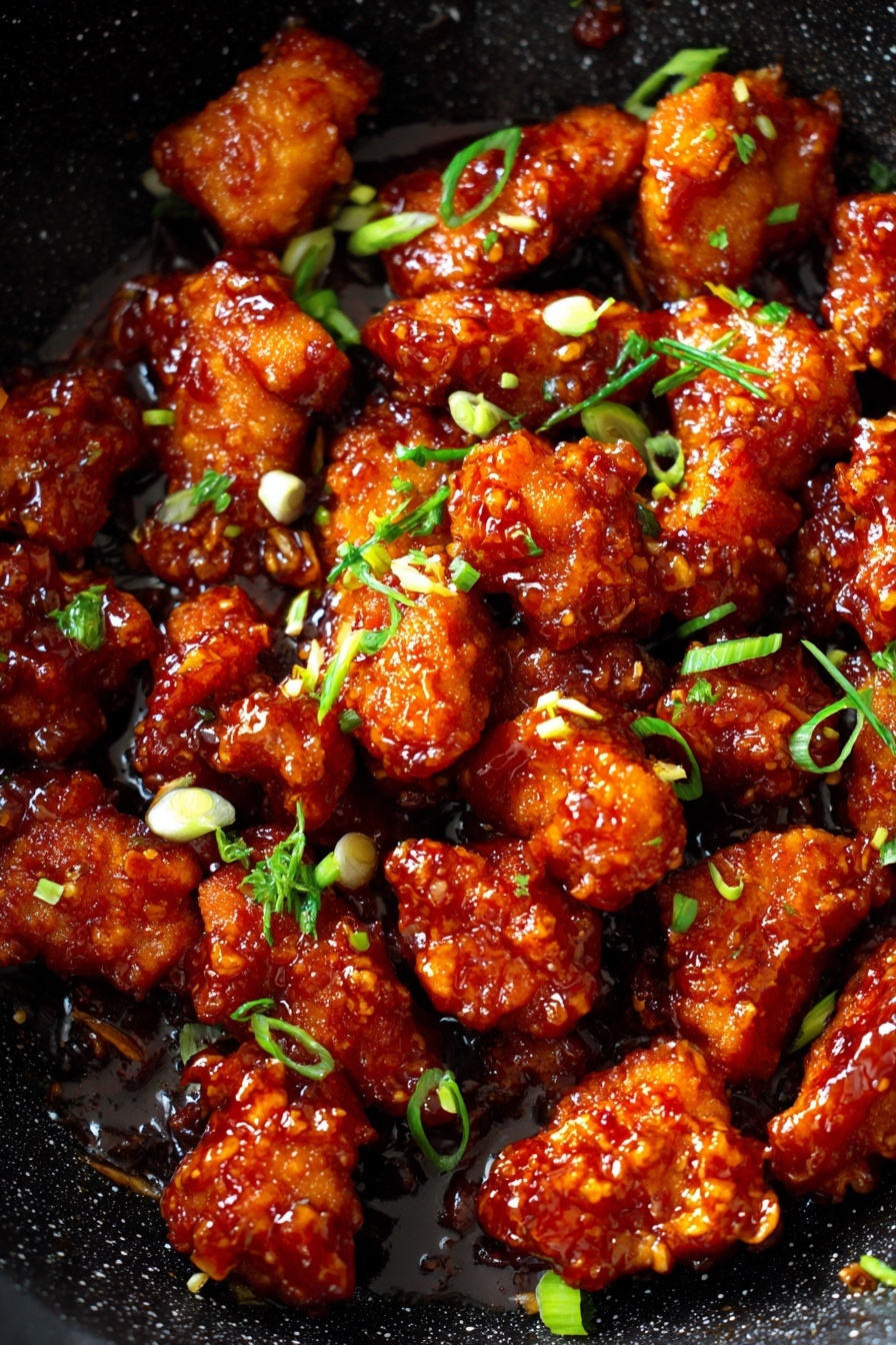This image shows many pieces of fried chicken coated with a shiny, reddish-brown sauce in a dark speckled pan. The chicken pieces look crispy under the sauce, with different shapes and sizes filling the pan. Small pieces of light green and white chopped green onions are scattered on top, adding color contrast. The pan has a rough texture and black color with little white dots. The overall look is warm and appetizing with juicy, glazed chicken pieces. Photo taken with an iphone --ar 2:3 --v 7 - Crispy Chilli Chicken, spicy crispy chicken recipe, Chinese-style chicken, easy fried chicken dishes, flavorful Asian chicken