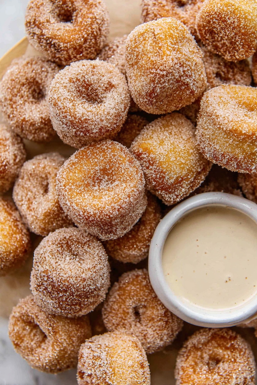 Cinnamon Sugar Pretzel Bites Recipe 6 The image shows many small, round donuts covered in sugar, piled closely together. The donuts have a golden-brown color with a rough sugar coating, giving them a sparkling look. In the center, there is a small, white bowl filled with a light creamy sauce. Everything is placed on a white marbled surface that adds a clean and bright background to the scene. The composition is close-up, focusing on the texture and color of the donuts and the bowl. photo taken with an iphone --ar 2:3 --v 7 - Cinnamon Sugar Pretzel Bites, easy pretzel bites, cinnamon sugar snack, homemade pretzel snacks, soft pretzel bites