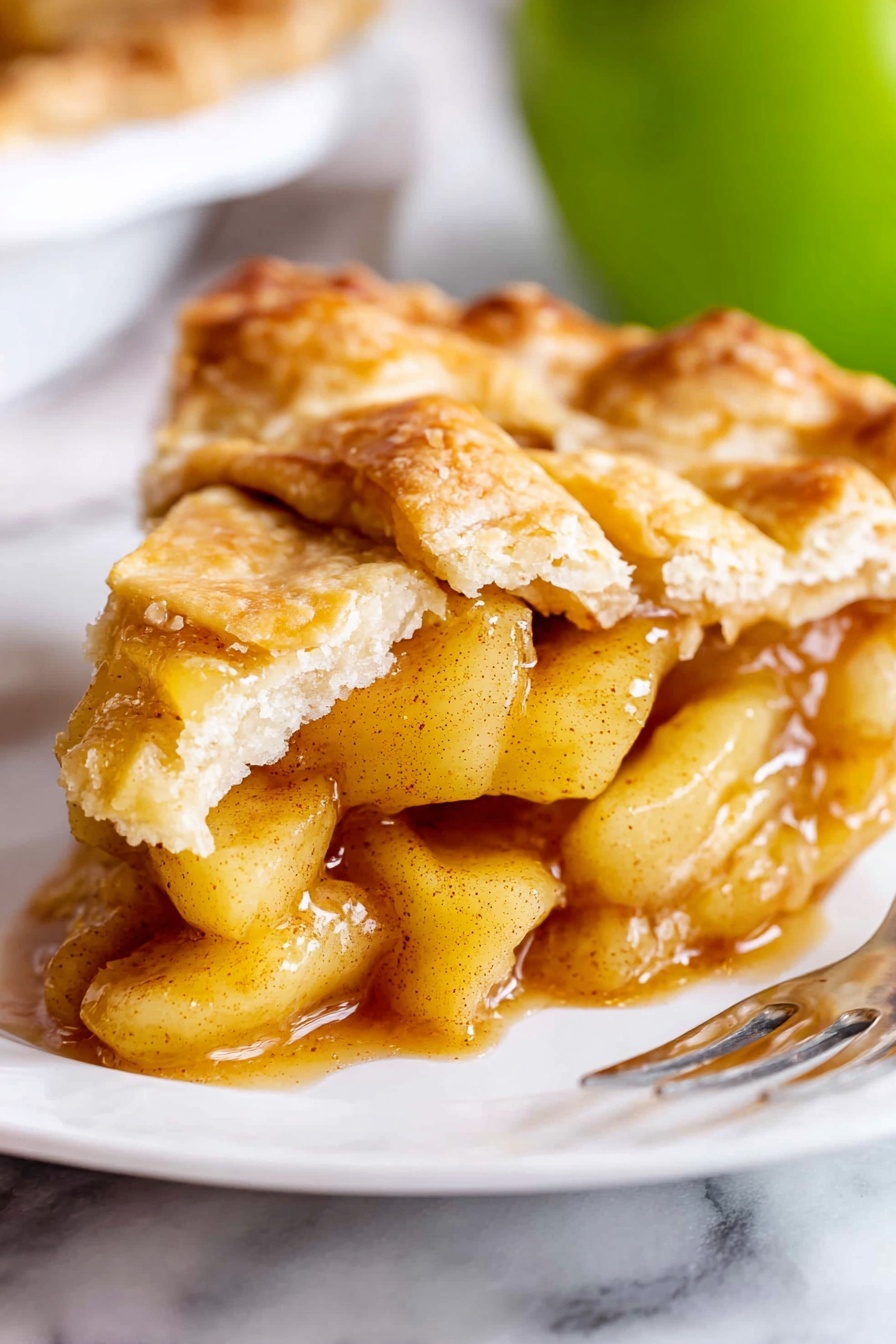 Perfect Apple Pie Recipe 6 A close-up of a slice of apple pie showing a flaky, golden-brown top crust with uneven, rustic edges. Beneath this crust, the filling has soft, light yellow apple chunks coated in a shiny, amber-colored sauce with specks of cinnamon. The pie rests on a white plate placed on a white marbled surface. In the background, there is a blurred green apple. A shiny silver fork is partially visible at the bottom right corner of the image. photo taken with an iphone --ar 2:3 --v 7 - Perfect Apple Pie, homemade apple pie, easiest apple pie recipe, flaky apple pie crust, caramelized apple filling