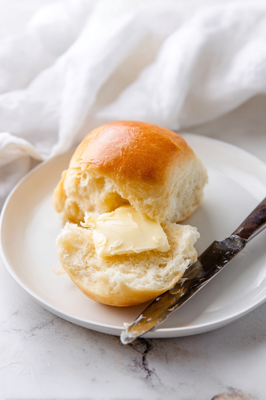 Fluffy Homemade Dinner Rolls Recipe 8 A white plate on a white marbled surface holds a soft, golden brown bread roll that is split open. The inside of the roll is light and fluffy with a smooth, shiny spread of butter melted into the upper half. Beside the roll lies an old-fashioned knife with a dark handle and a thick, creamy layer of butter on its blade. In the background, a white cloth is softly draped, adding a cozy feel to the scene. photo taken with an iphone --ar 2:3 --v 7 - Fluffy Homemade Dinner Rolls, soft dinner rolls, easy bread recipe, homemade dinner rolls, perfect dinner rolls