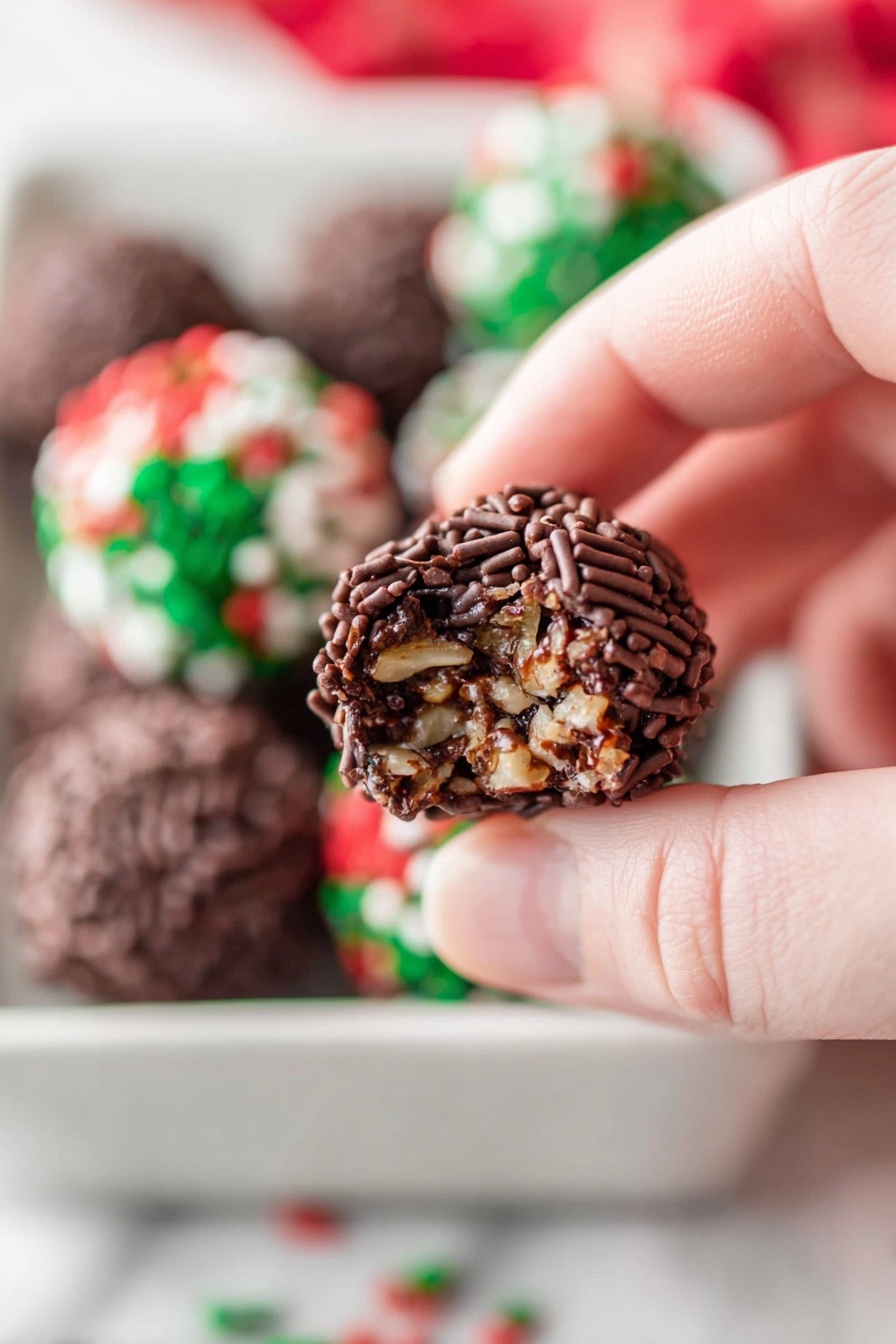 No Bake Chocolate Rum Balls Recipe 6 A close-up image of a woman's hand holding a small chocolate ball covered in dark brown chocolate sprinkles, with a bite taken out showing a dense, textured inside of mixed nuts and chocolate. In the blurred background, more chocolate balls with red, green, and white sprinkles are visible, all placed in a white tray on a white marbled surface. Photo taken with an iphone --ar 2:3 --v 7 - No Bake Chocolate Rum Balls, easy rum ball recipes, holiday no-bake desserts, chocolate holiday treats, boozy chocolate bites