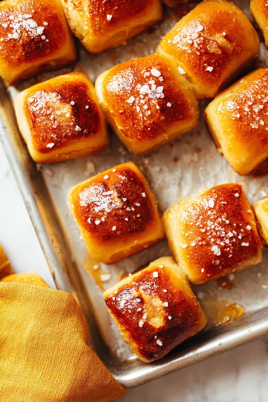 Buttery Soft Pretzel Bites Recipe 6 The image shows a metal baking tray holding about a dozen small square pretzel bites. Each piece has a golden brown top with a slightly darker toasted patch in the center and is sprinkled with coarse salt crystals. The background is a white marbled texture, and a mustard-yellow cloth is partially visible in the bottom left corner. The pretzel bites are neatly spaced, showing a soft and fluffy texture with a shiny crust. Photo taken with an iphone --ar 2:3 --v 7 - Buttery Soft Pretzel Bites, easy pretzel bites recipe, homemade pretzel snacks, soft pretzel bites how to, chewy buttery pretzels