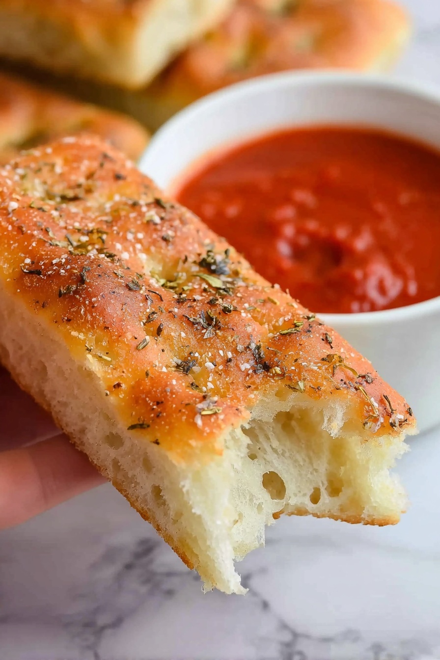 Copycat Pizza Hut Breadsticks Recipe 6 A close-up of a woman's hand holding a rectangular piece of focaccia bread with a light golden crust on top sprinkled with coarse salt and dried herbs, showing the soft, airy white inside at the bitten end. In the blurry background, a white bowl filled with thick red marinara sauce is visible, alongside another piece of focaccia bread partly visible on a white marbled surface. photo taken with an iphone --ar 2:3 --v 7 - Copycat Pizza Hut Breadsticks, homemade breadsticks, garlic butter breadsticks, soft cheesy breadsticks, easy breadstick recipe