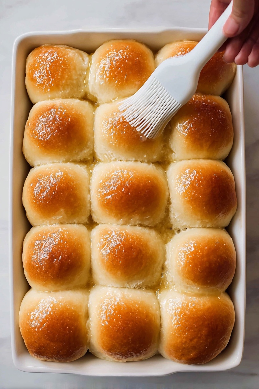 Fluffy Homemade Dinner Rolls Recipe 6 A white baking dish holds fifteen soft, fluffy golden brown dinner rolls arranged in a 3 by 5 grid. Each roll has a shiny, smooth top with a slightly darker brown in the middle and a light fluffy texture on the sides where they touch. A white silicone brush held by a woman's hand gently spreads butter or glaze on the top of one roll near the center. The background is a white marbled texture. photo taken with an iphone --ar 2:3 --v 7 - Fluffy Homemade Dinner Rolls, soft dinner rolls, easy bread recipe, homemade dinner rolls, perfect dinner rolls