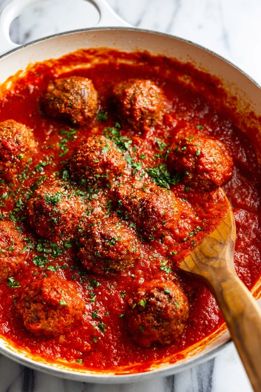 Cranberry Meatballs Recipe 8 The image shows a close-up of meatballs covered in thick red tomato sauce inside a white pan. The sauce is rich and smooth with a glossy texture, and the meatballs are round with a slightly crispy surface peeking through the sauce layer. Bright green chopped herbs are sprinkled evenly over the top, adding a fresh pop of color. A wooden spatula is partially visible on the right side, touching the sauce and meatballs. The background is a white marbled texture. photo taken with an iphone --ar 2:3 --v 7 - Cranberry Meatballs, holiday meatballs with cranberry sauce, easy cranberry meatballs, savory cranberry meatball recipe, festive appetizer ideas