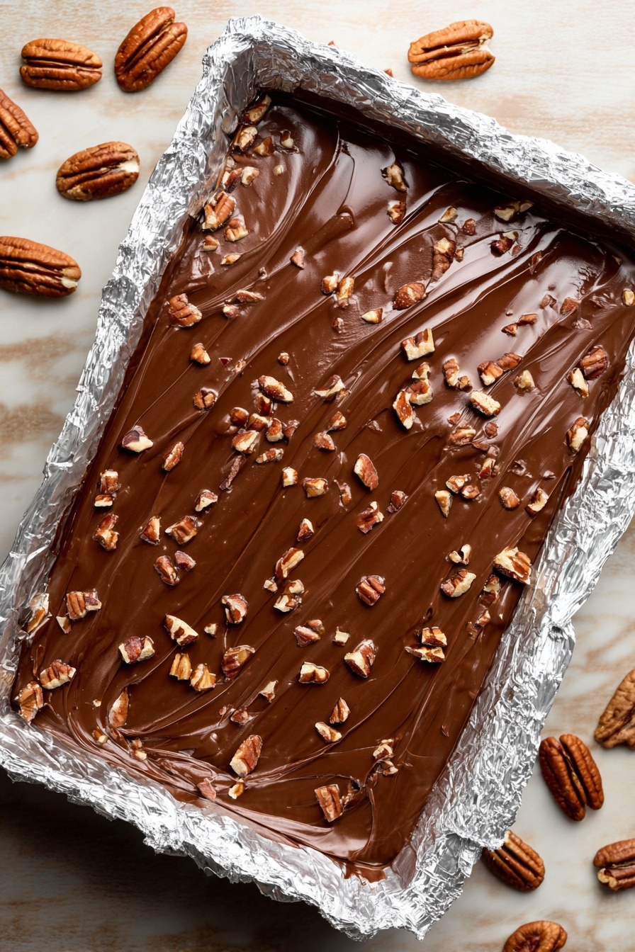 The image shows a rectangular foil-lined pan filled with a rich, smooth chocolate layer that has a glossy texture and visible swirl lines on the surface. On top of the chocolate layer, there are many small pieces of chopped pecans scattered evenly. The pan is placed on a white marbled texture with several whole and halved pecans around it. photo taken with an iphone --ar 2:3 --v 7 - Saltine Toffee Christmas Crack, holiday dessert, easy Christmas treats, salty and sweet snacks, no-bake holiday sweets