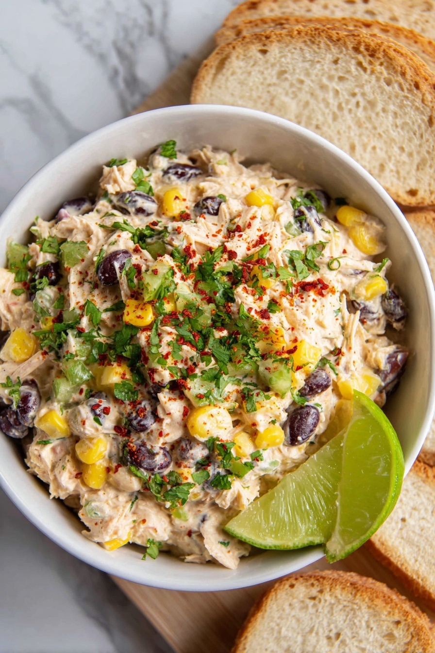 Southwest Chicken Salad Recipe 6 A white bowl filled with a layered salad mix sits on a white marbled surface with slices of light bread arranged diagonally next to it. The salad has a creamy layer with light beige shredded pieces, black beans, yellow corn kernels, small red bits, and green slices, topped with chopped fresh green herbs and a sprinkle of red seasoning. A bright green lime wedge rests on the right side of the bowl edge. The overall look is fresh with a mix of creamy, colorful, and textured ingredients. Photo taken with an iphone --ar 2:3 --v 7 - Southwest Chicken Salad, Southwest Chicken Salad Ingredients, Southwest Chicken Salad Ideas, Southwest Chicken Salad Healthy, Easy Southwest Chicken Salad