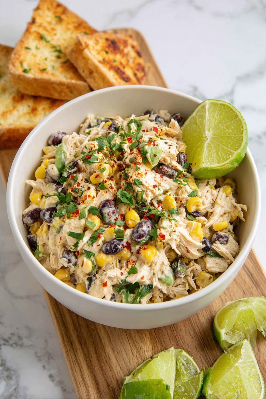 Southwest Chicken Salad Recipe 8 A white bowl filled with a creamy salad made of shredded chicken mixed with yellow corn kernels, black beans, small pieces of green and red vegetables, and topped with chopped green herbs and a sprinkle of red spice. A green lime wedge is placed inside the bowl on one side. The bowl sits on a wooden board next to more lime wedges and toasted white bread triangles, all on a white marbled surface. photo taken with an iphone --ar 2:3 --v 7 - Southwest Chicken Salad, Southwest Chicken Salad Ingredients, Southwest Chicken Salad Ideas, Southwest Chicken Salad Healthy, Easy Southwest Chicken Salad